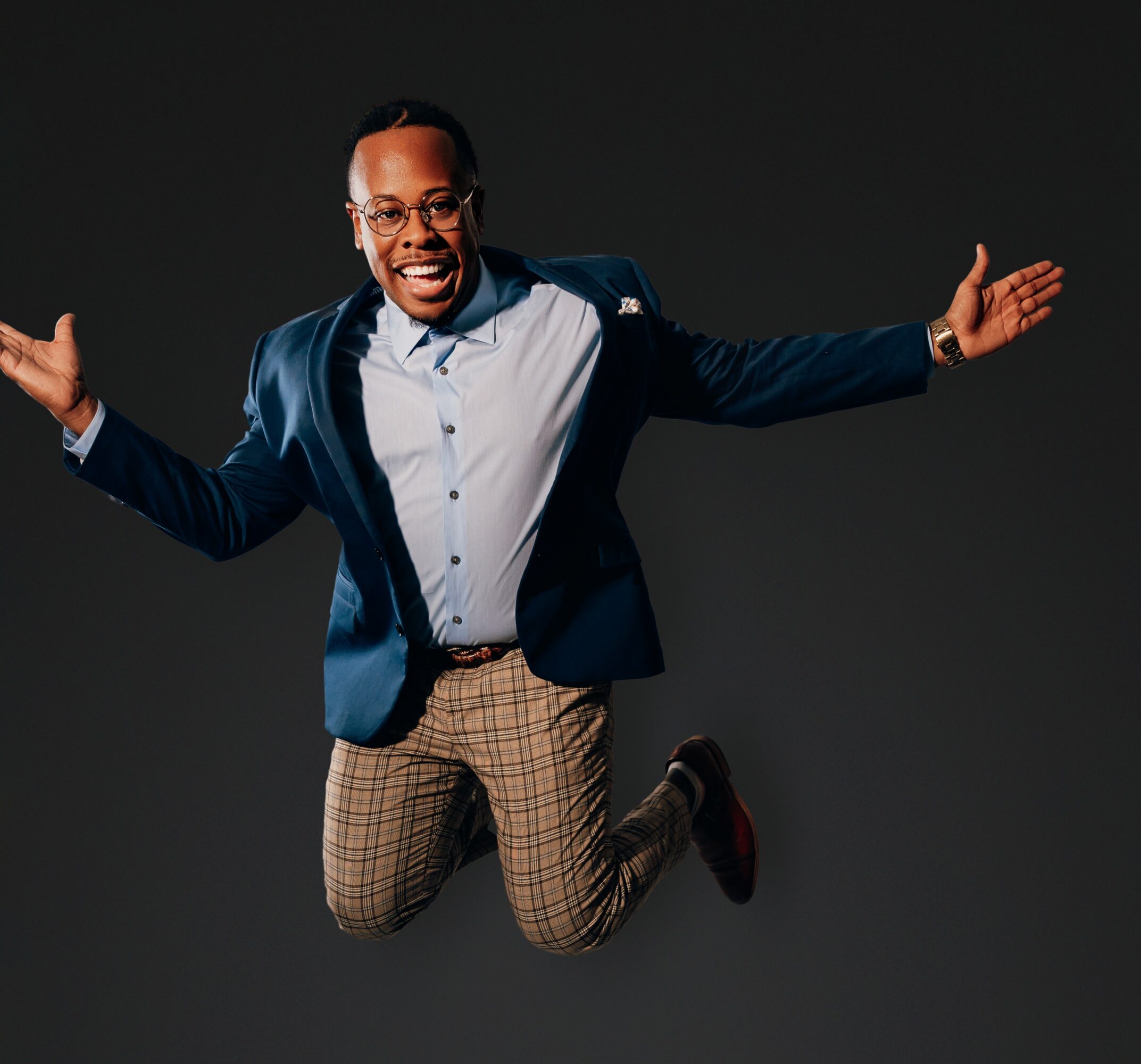 Person jumping with arms outstretched, smiling, wearing glasses, a blue blazer, light shirt, and checkered pants, against a dark background.