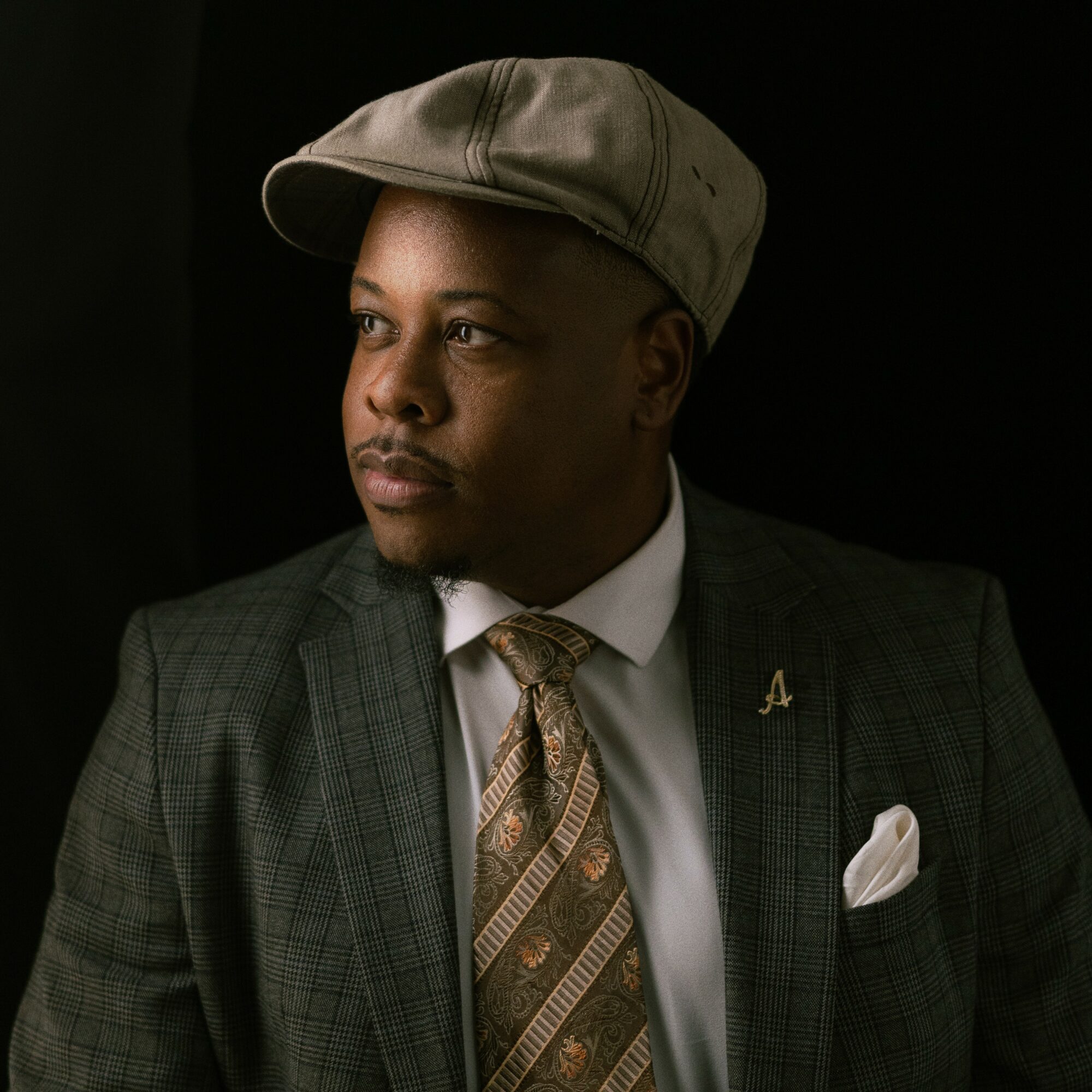 Man wearing a gray cap, suit, and patterned tie, looking to the side against a dark background.