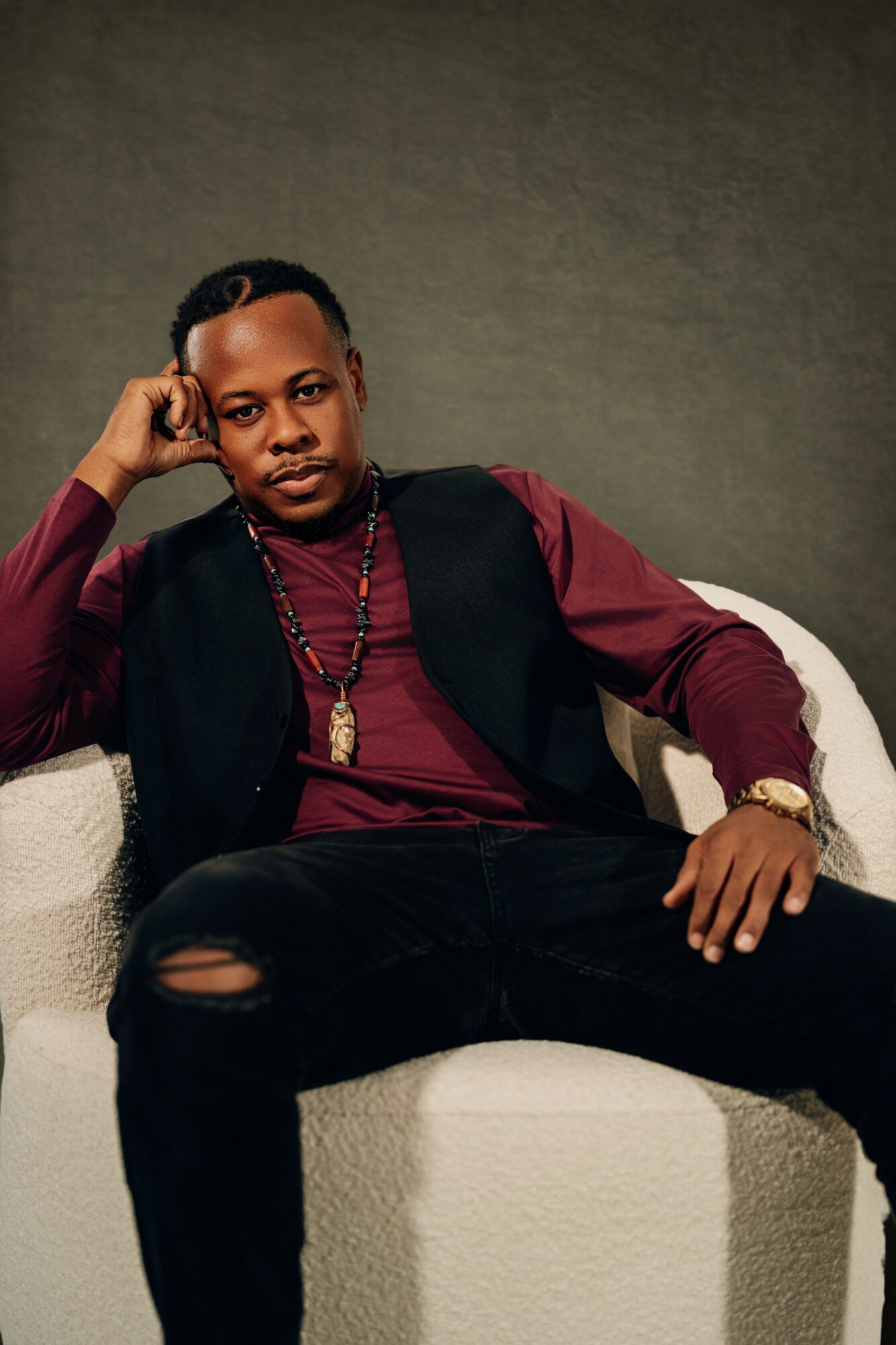 Man sitting on a white couch, resting his head on his hand, wearing a maroon shirt, black vest, and gold jewelry.
