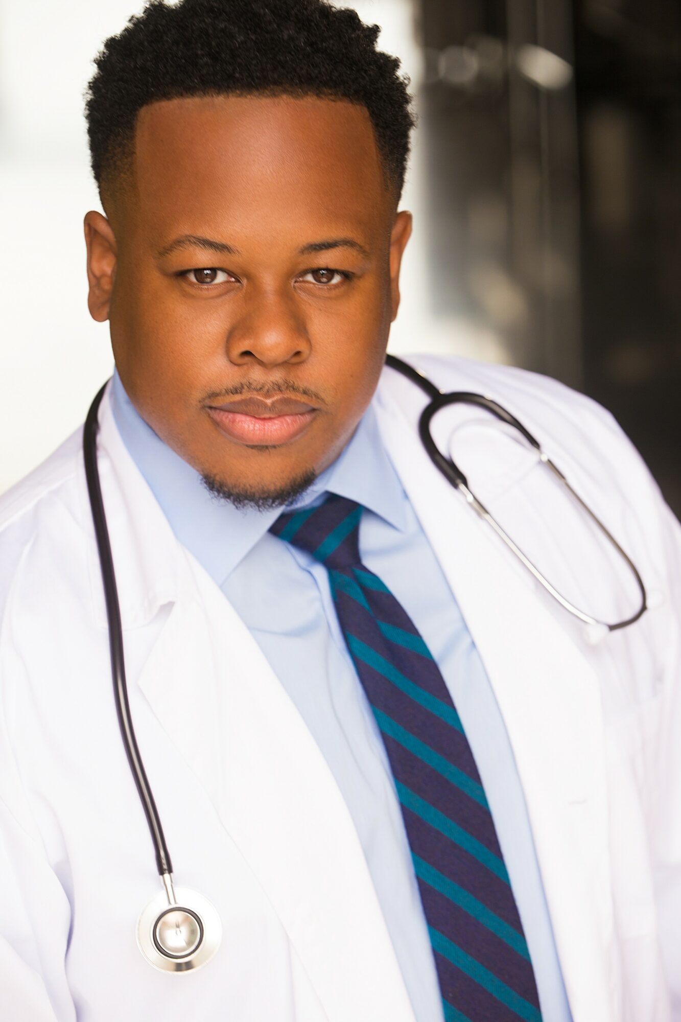 Male doctor in white coat with stethoscope, wearing blue shirt and striped tie, looking at camera.