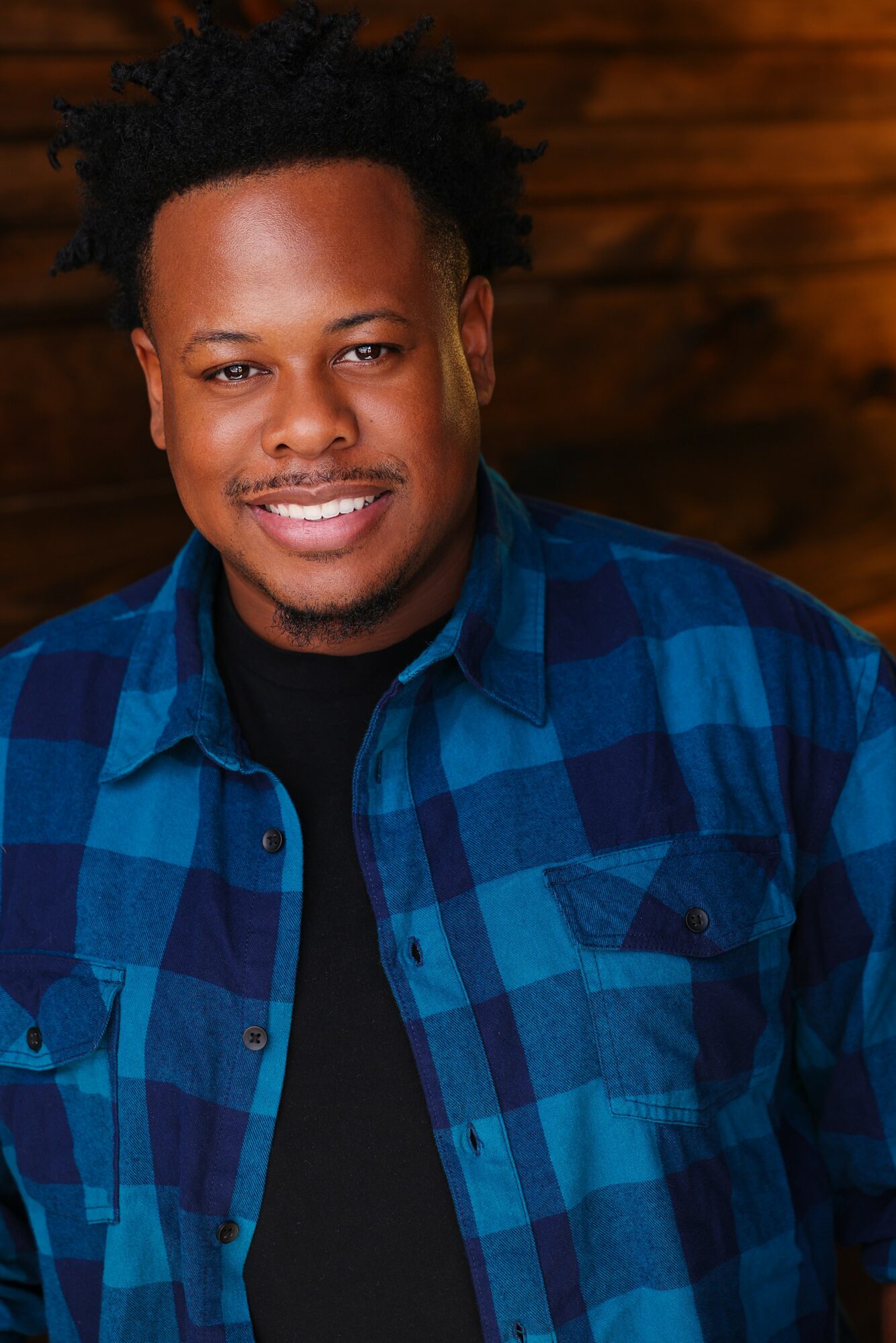 Portrait of a smiling man with curly hair wearing a blue plaid shirt and black t-shirt, wooden background