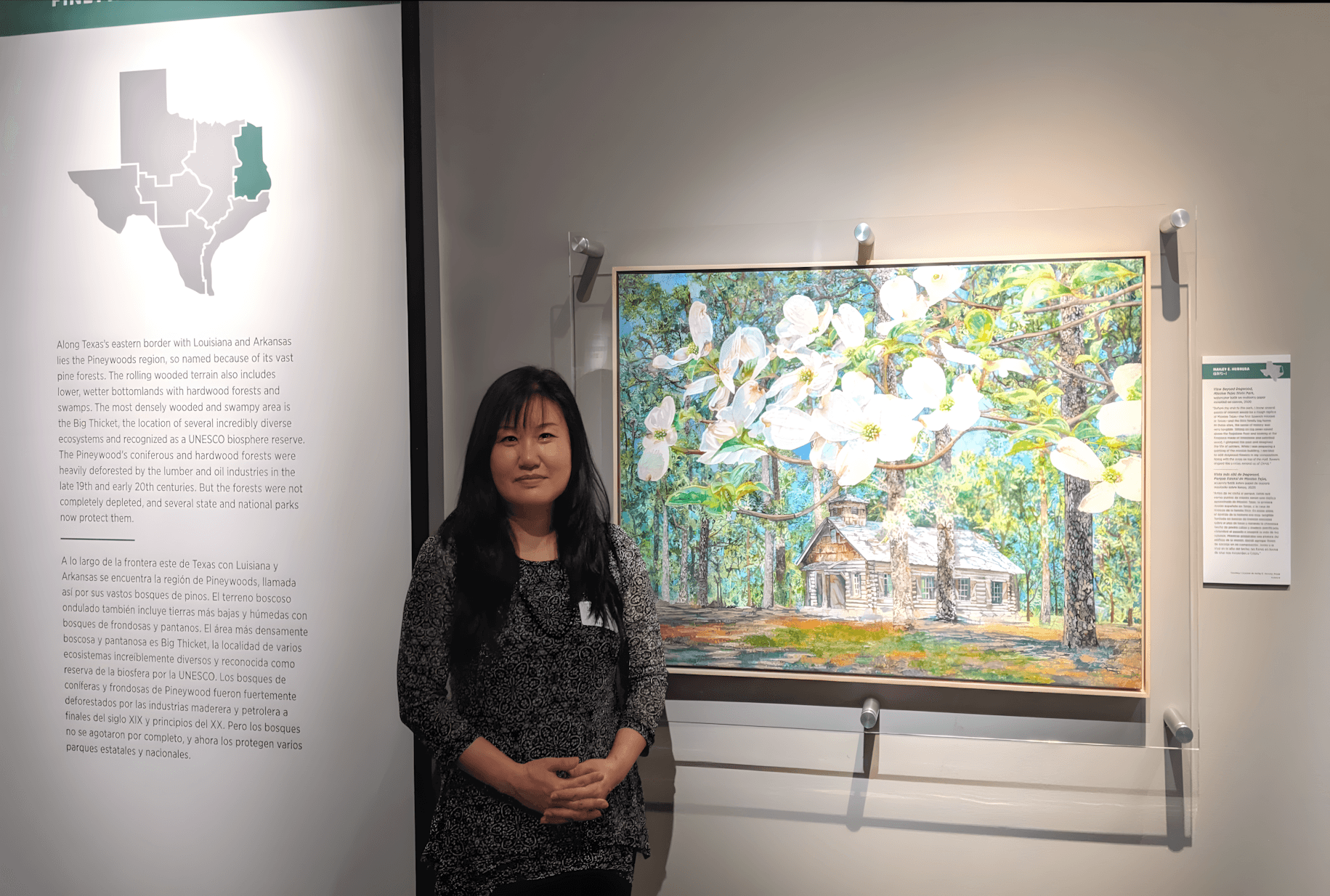 Woman standing next to a colorful landscape painting of a house surrounded by trees in an art gallery.