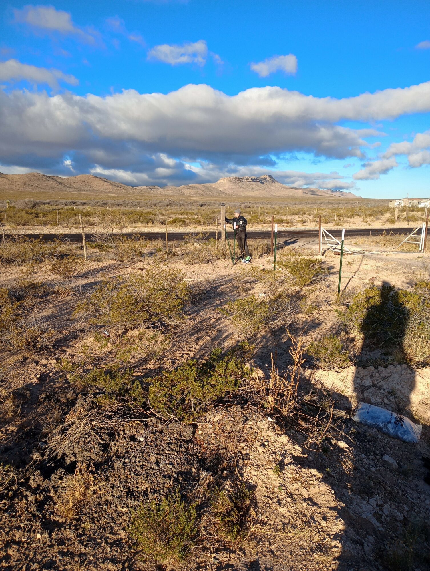 Landscape with a rocky hillside, sparse vegetation, and a partly cloudy sky, with power lines and a person in the distance.