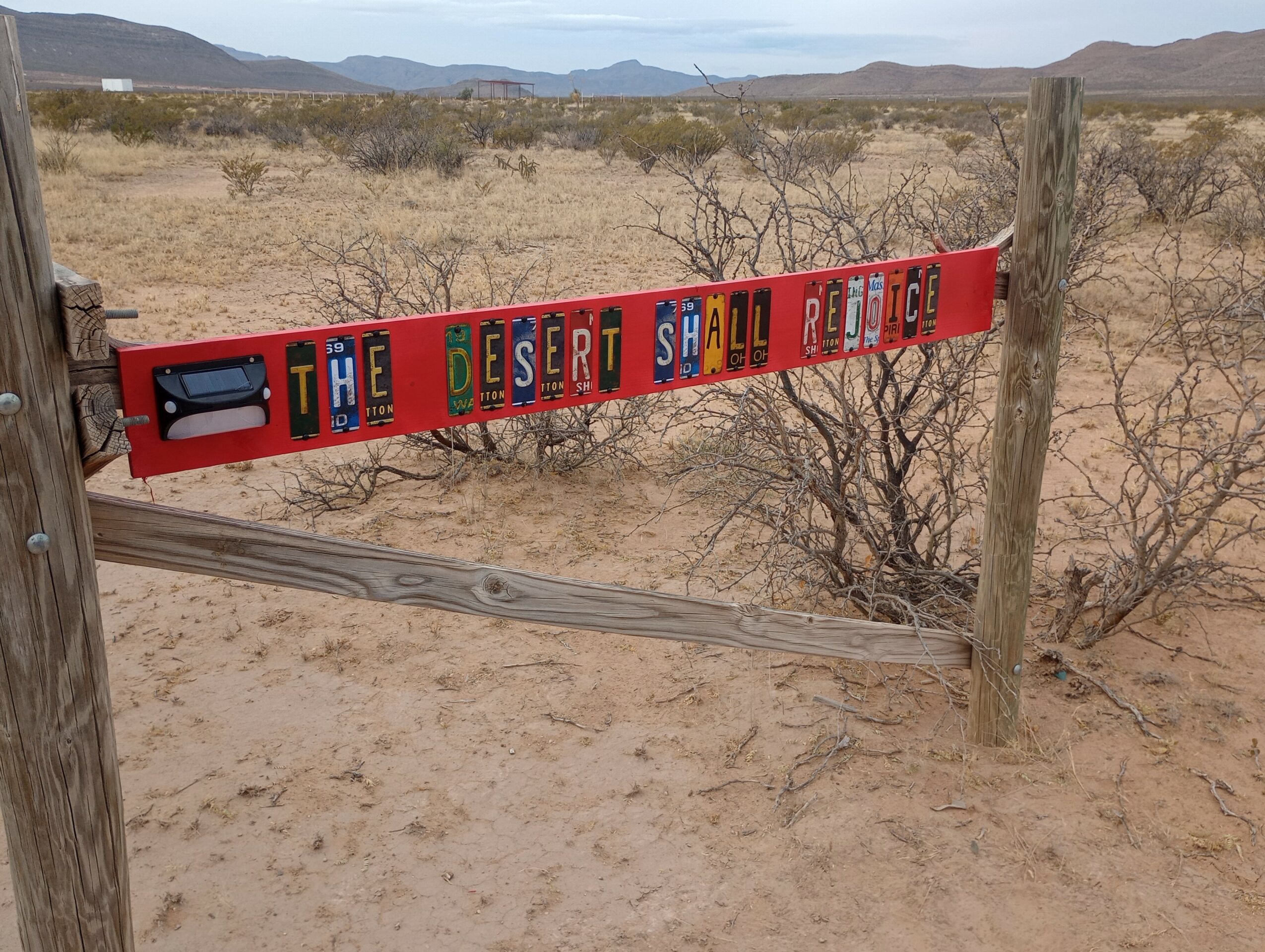 Desert landscape with a sign reading 'The Desert Shall Rejoice' in colorful letters, dry bushes, and mountains in the background.