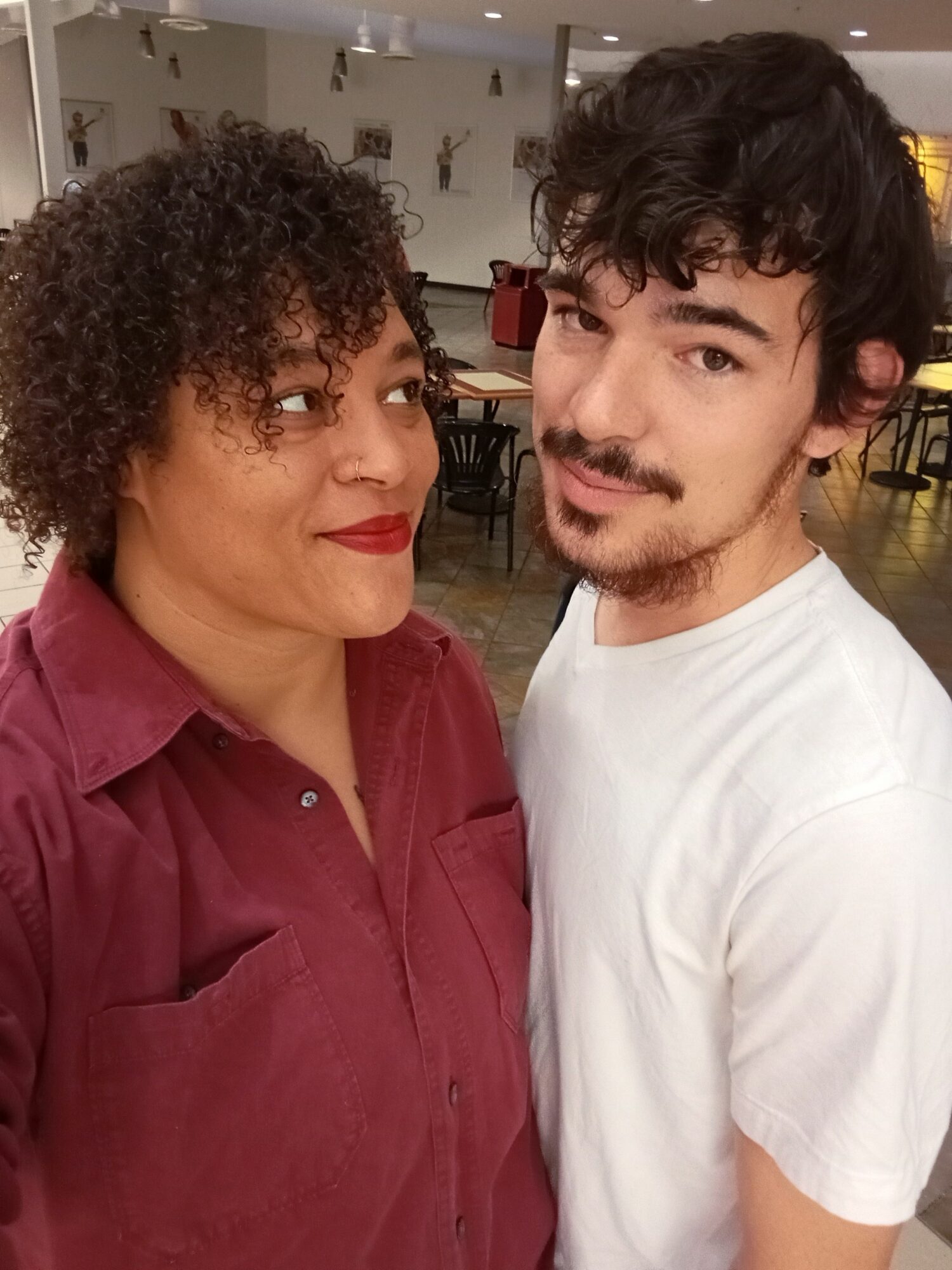 Two people, a woman with curly hair and a man with dark hair and a beard, pose together indoors.