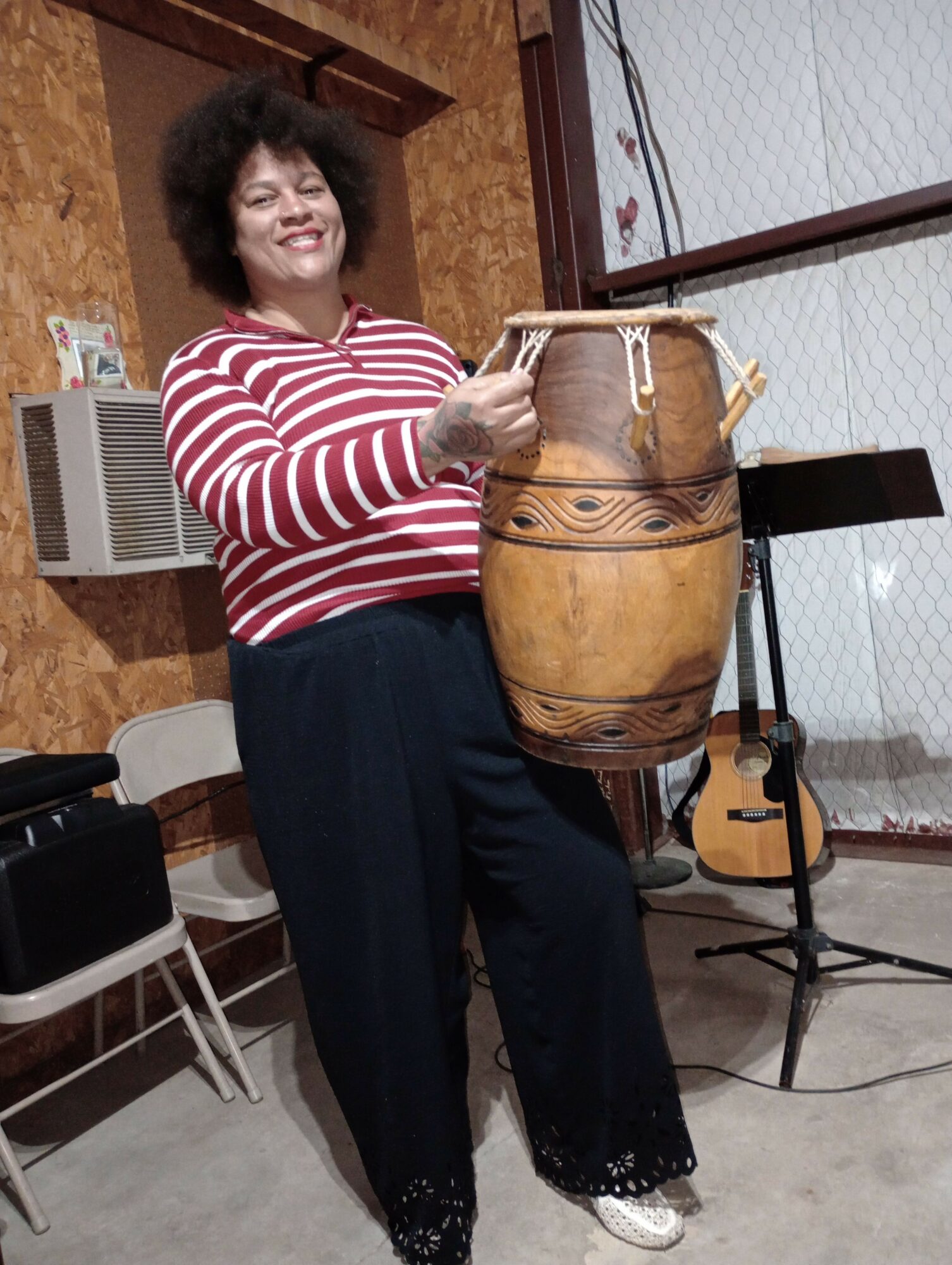Woman in striped shirt and black pants standing next to a large drum, guitar, and microphone stand in a room.