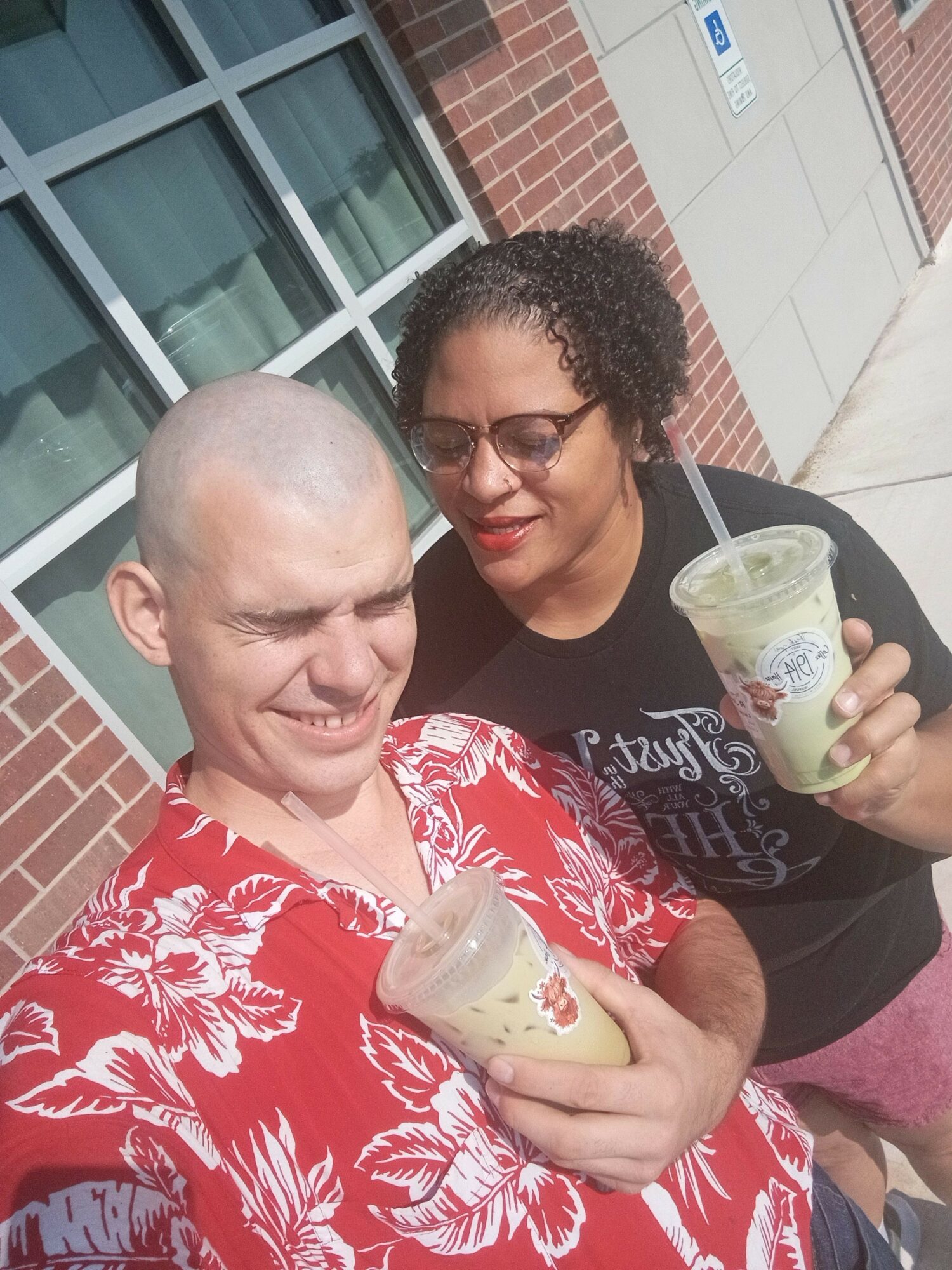 Two people smiling, holding drinks, standing outside a building with large windows and brick wall.