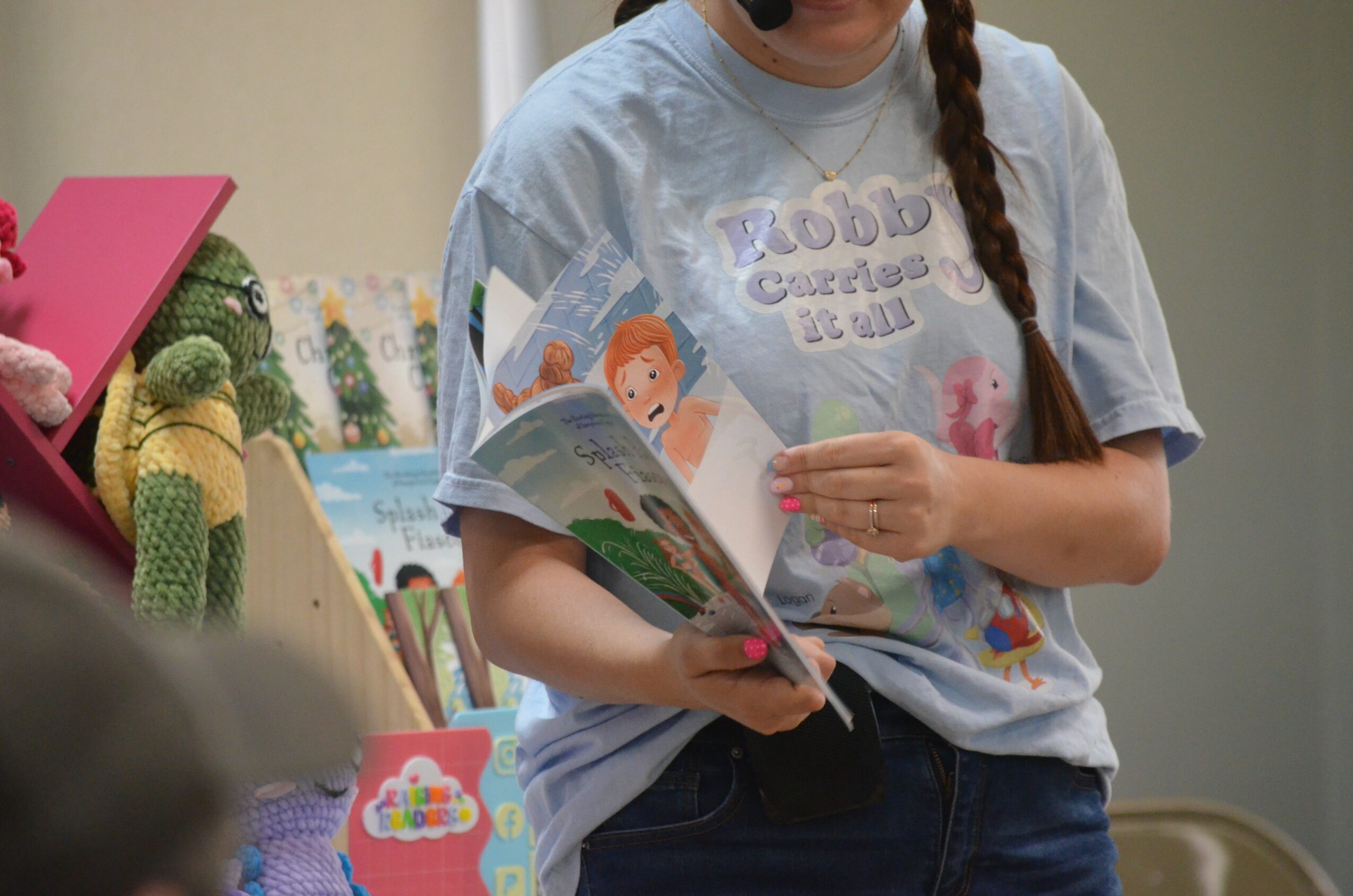 Person holding a colorful children's book, wearing a light gray T-shirt with a cartoon design, in an indoor setting.