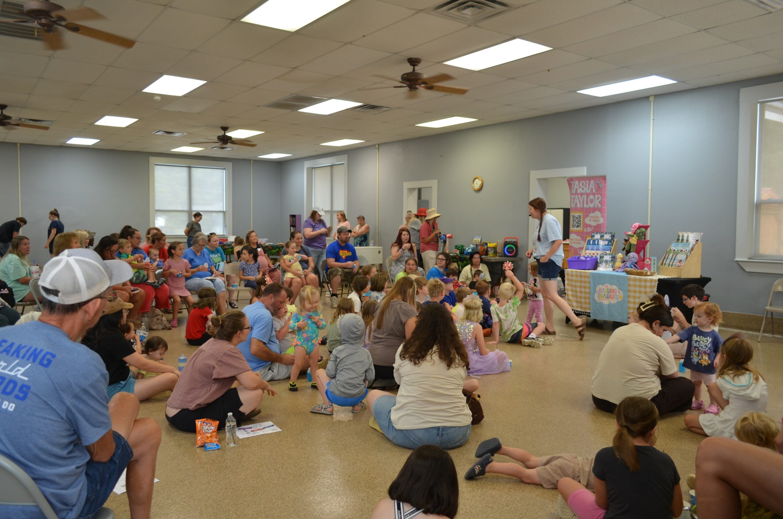 Large group of children and adults gathered in a room, sitting on the floor, with tables and displays at the front.