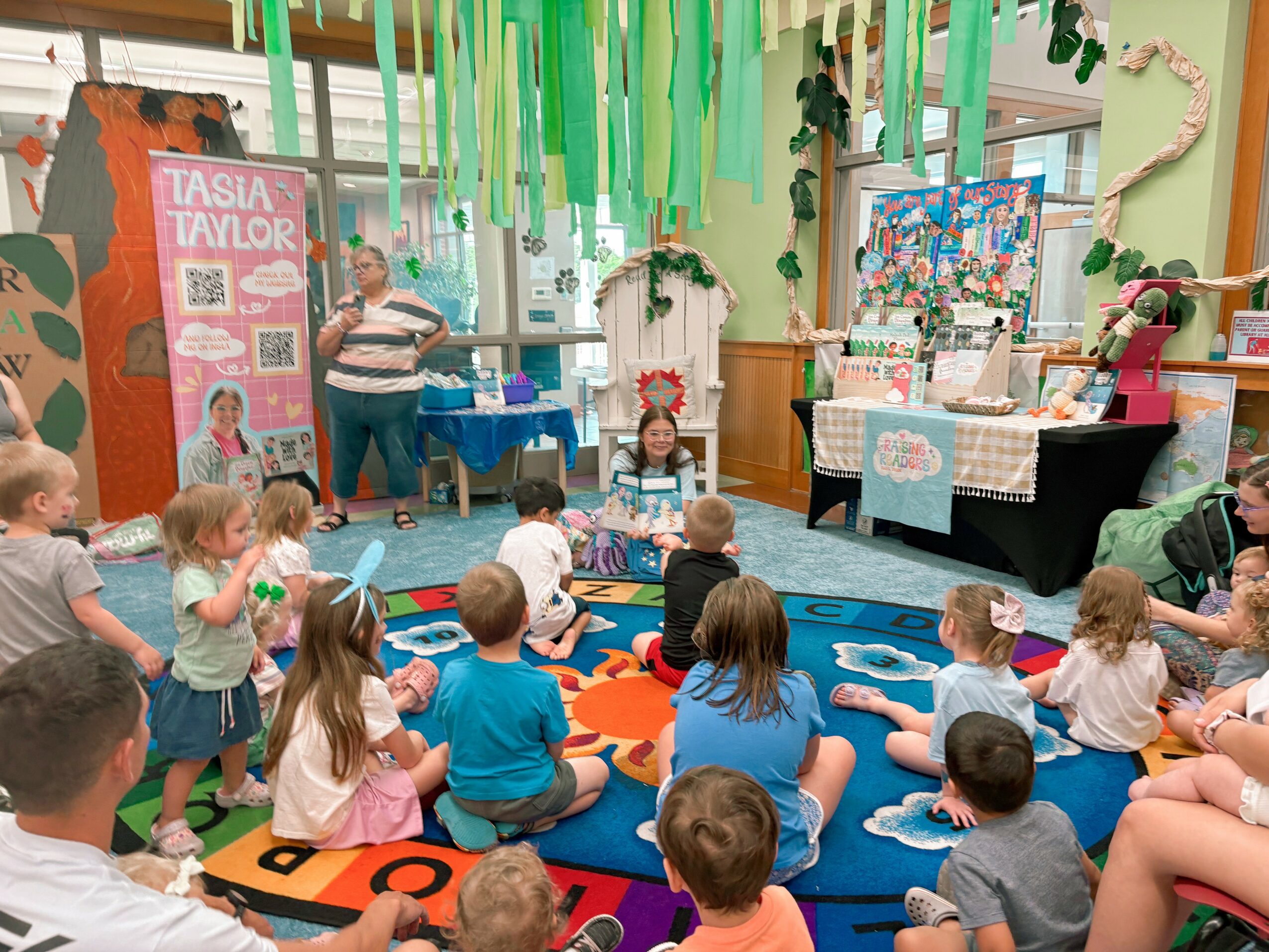 Children sitting on a colorful carpet watching a woman perform in a decorated indoor space with green streamers and display tables.