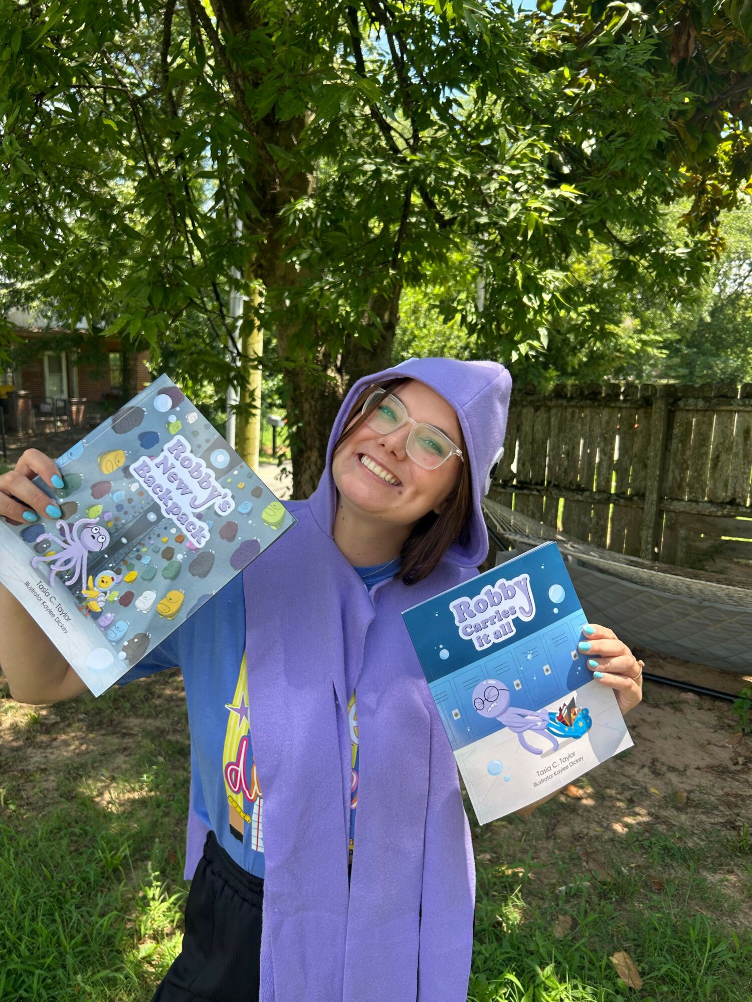 Person wearing a purple raincoat holding two colorful books outdoors, smiling, with trees and a wooden fence in the background.