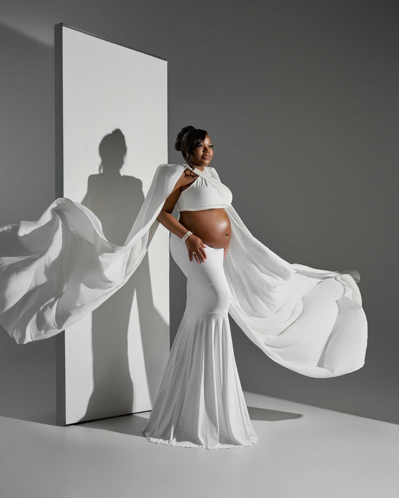 Pregnant woman in white dress with flowing fabric, standing against a gray background with shadow, holding her belly.