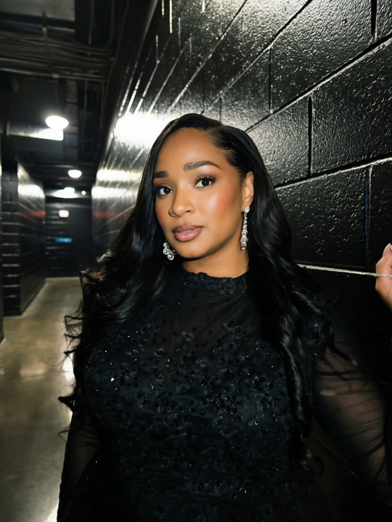 Young woman with long black hair, wearing earrings and a black dress, standing in a dark corridor with black brick walls.