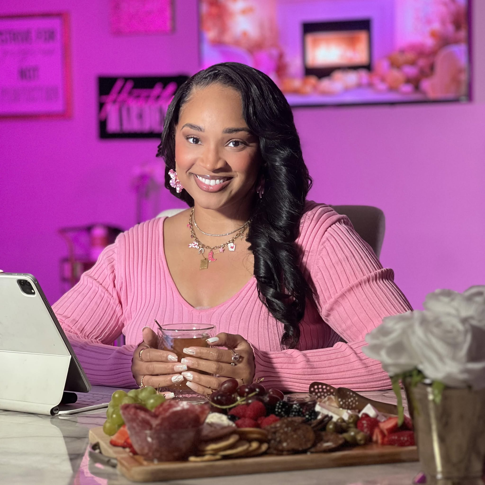 Smiling woman with long black hair, wearing pink sweater, sitting at desk with food and tablet, pink-lit background.