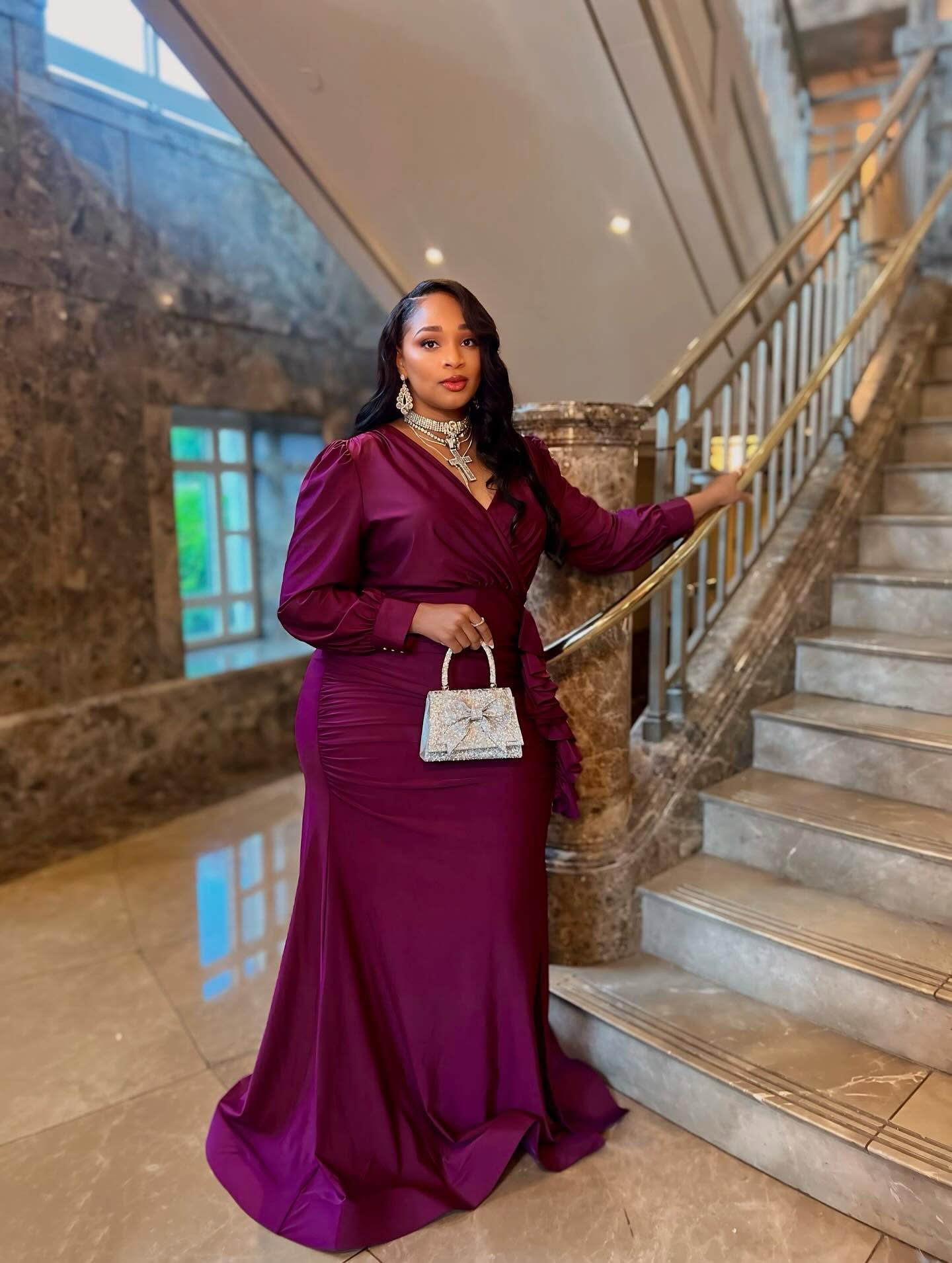 Woman in a long, elegant purple gown holding a small handbag, standing beside a staircase in a luxurious interior.