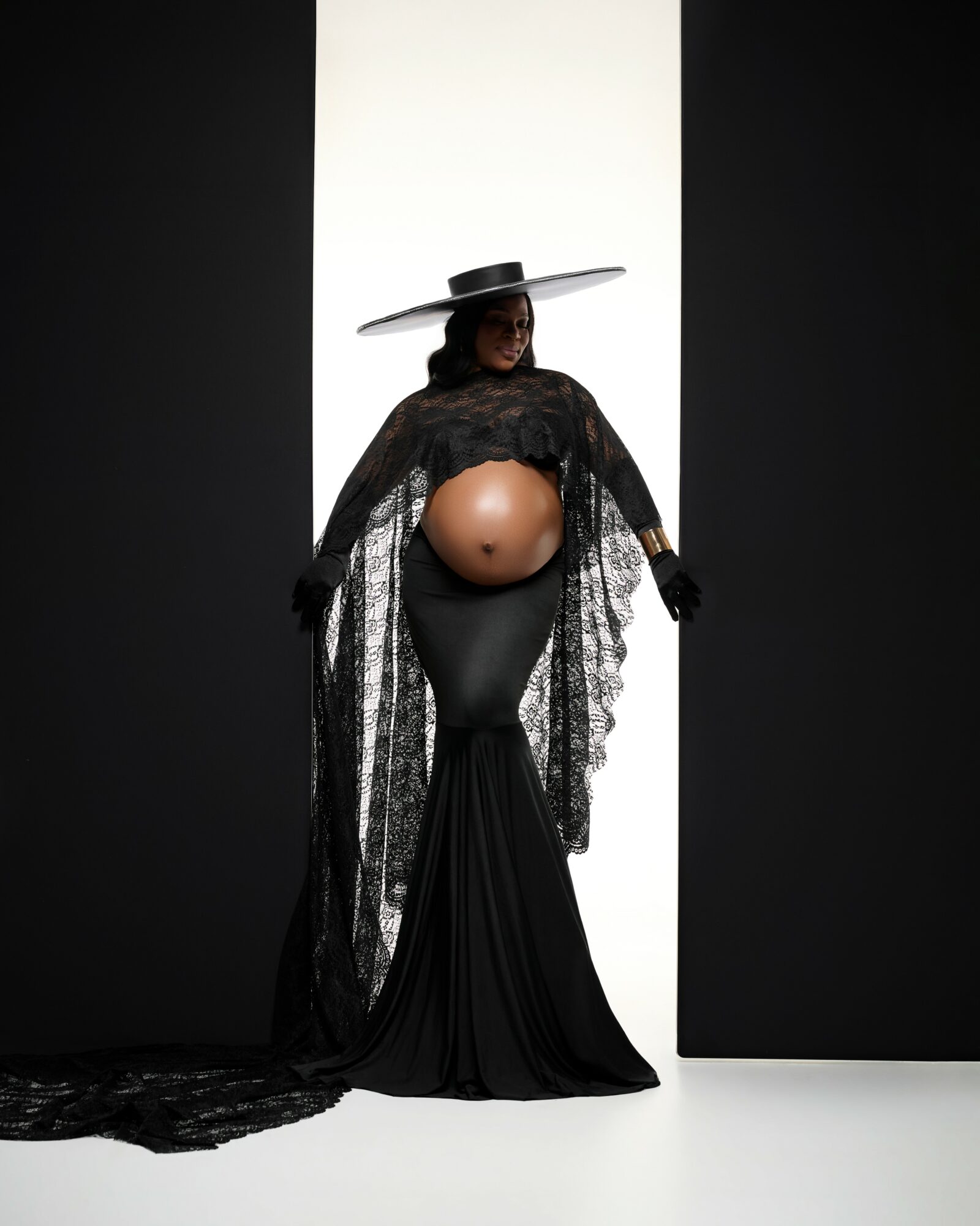 Person wearing a large hat, black lace cape, and black dress with a prominent pregnant belly, standing against a black and white background.