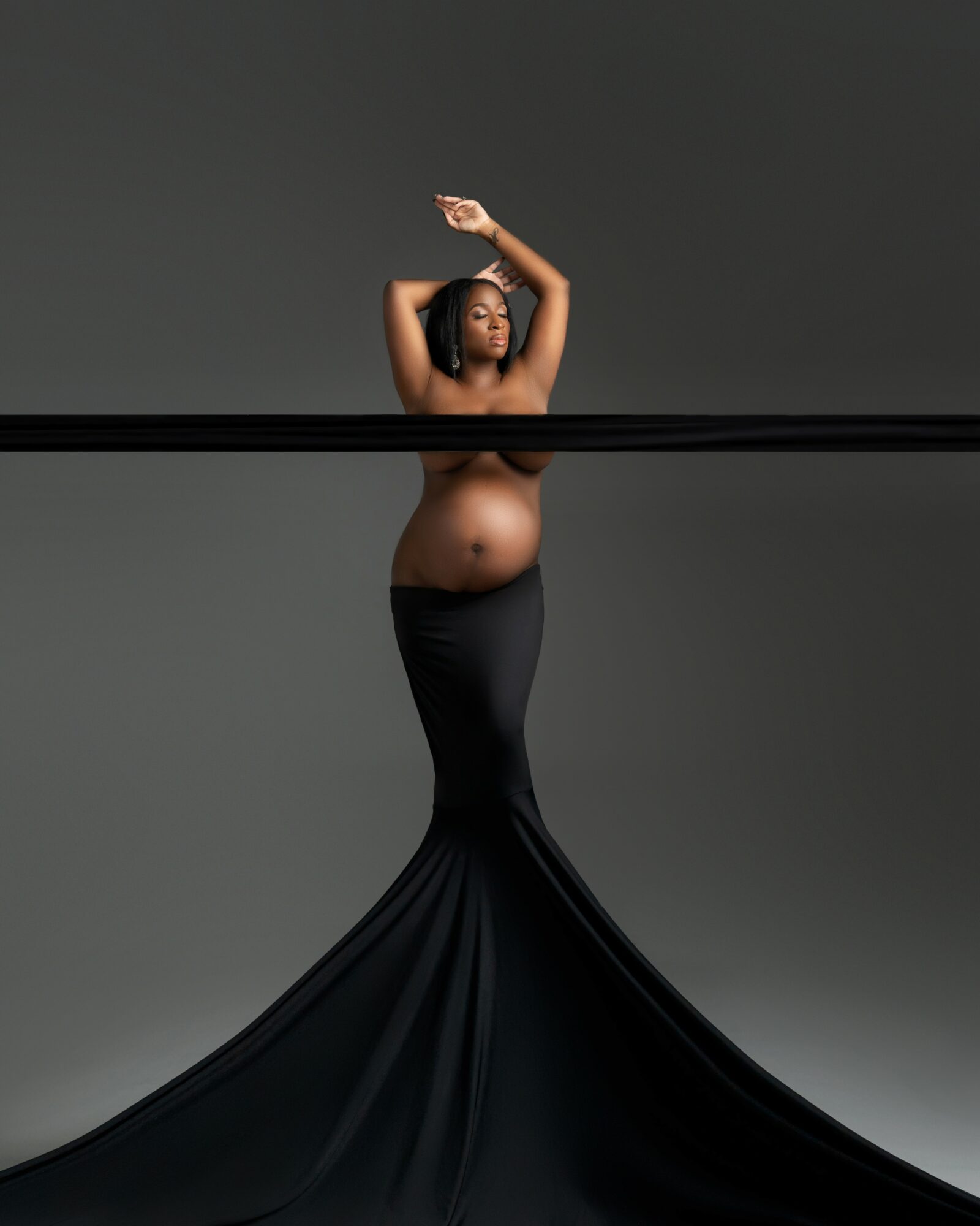 Pregnant woman with arms raised, wearing a long black skirt, standing against a gray background.
