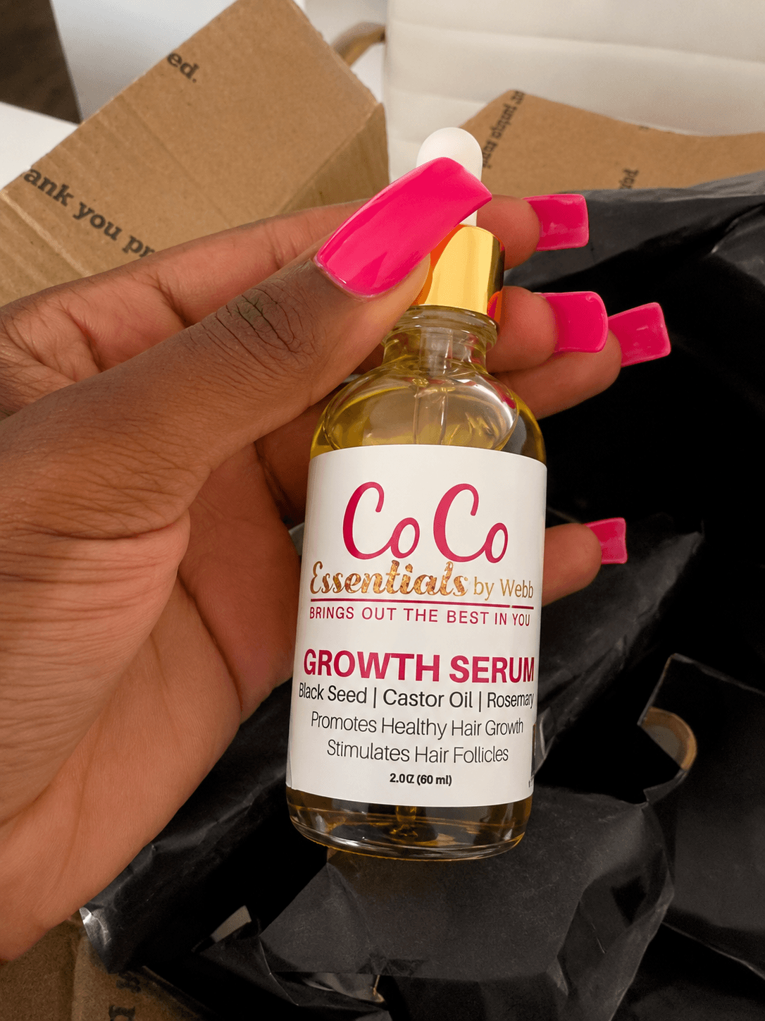 Hand holding a small bottle of Co Co Essentials Growth Serum with pink nails, background includes cardboard and black packaging.