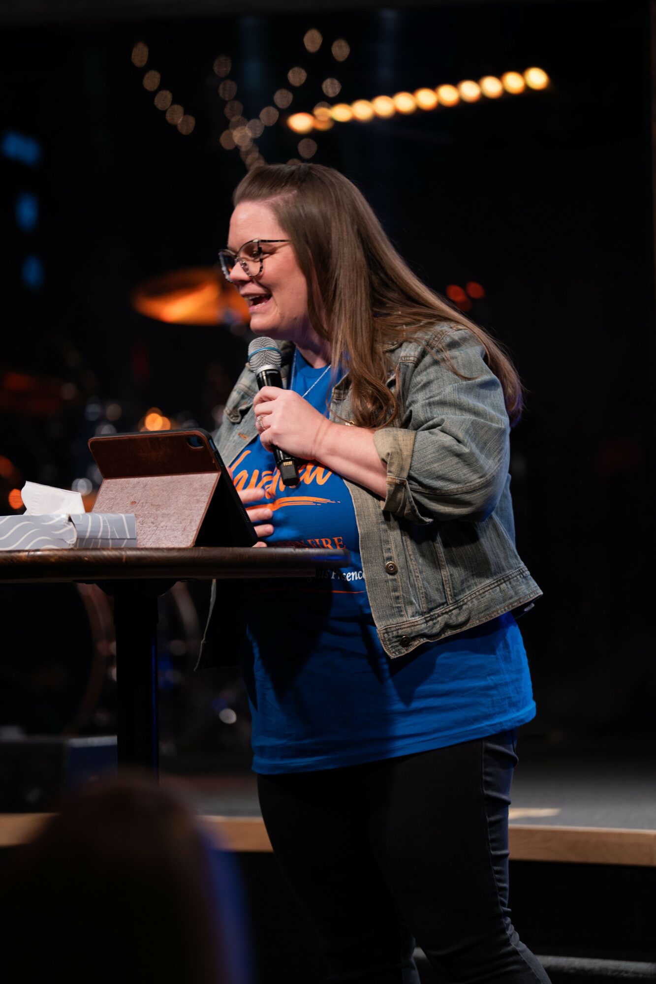 Young woman with glasses, holding a microphone, smiling, standing at a table on stage with blurred lights in background.