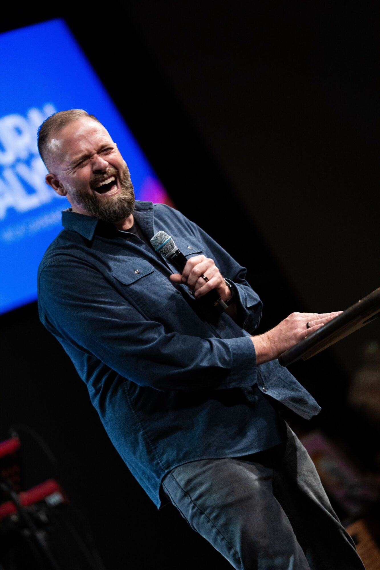 Man with a beard laughing and holding a microphone on stage, with a blue screen in the background.