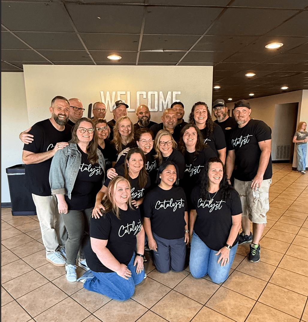 Group of people smiling and posing indoors in front of a white wall with 'WELCOME' text, some wearing matching black shirts.