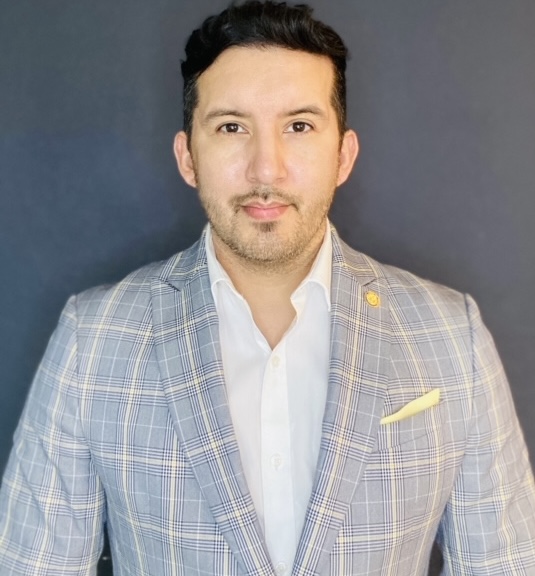 Portrait of a man with dark hair, wearing a light plaid suit and white shirt, against a dark background.