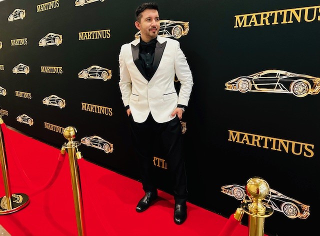 Man in a white tuxedo jacket and black pants standing on a red carpet with a black backdrop featuring car illustrations and the word 'MARTINUS'.