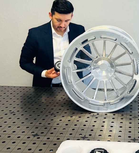 Man in suit examining a large metallic wheel on a perforated table, plain wall background.