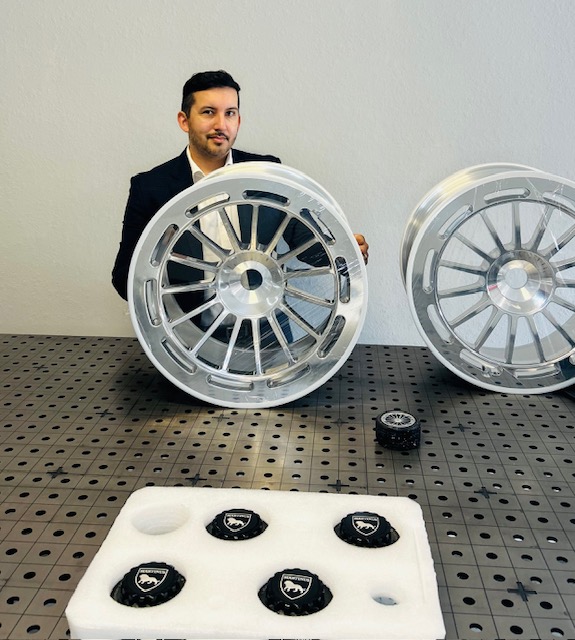 Man in a suit stands behind two large wheel rims on a table, with a small object in front of them, against a plain wall.