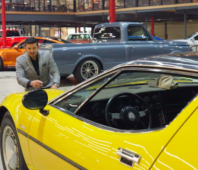 Man in gray suit leaning on yellow sports car inside a showroom with classic cars in background.