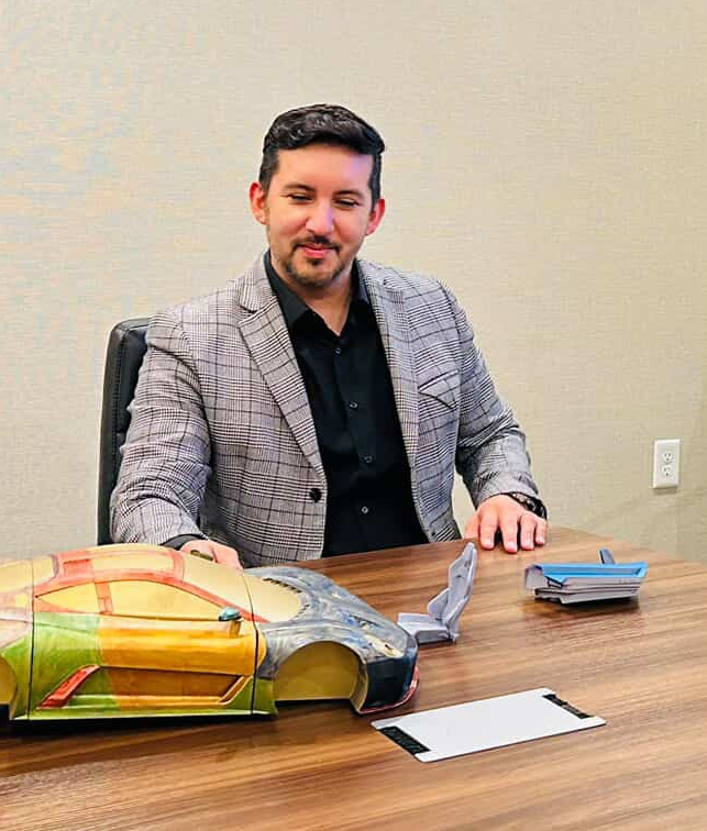 Man in a checkered blazer sitting at a table with a colorful bag and papers, smiling, against a plain wall.