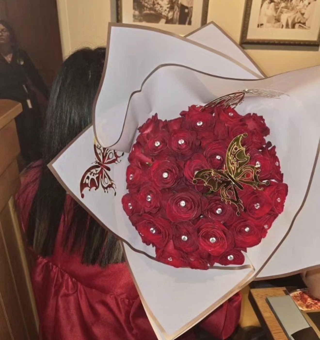 Person holding a bouquet of red roses with decorative butterfly and pearl accents, indoors with framed pictures on the wall.