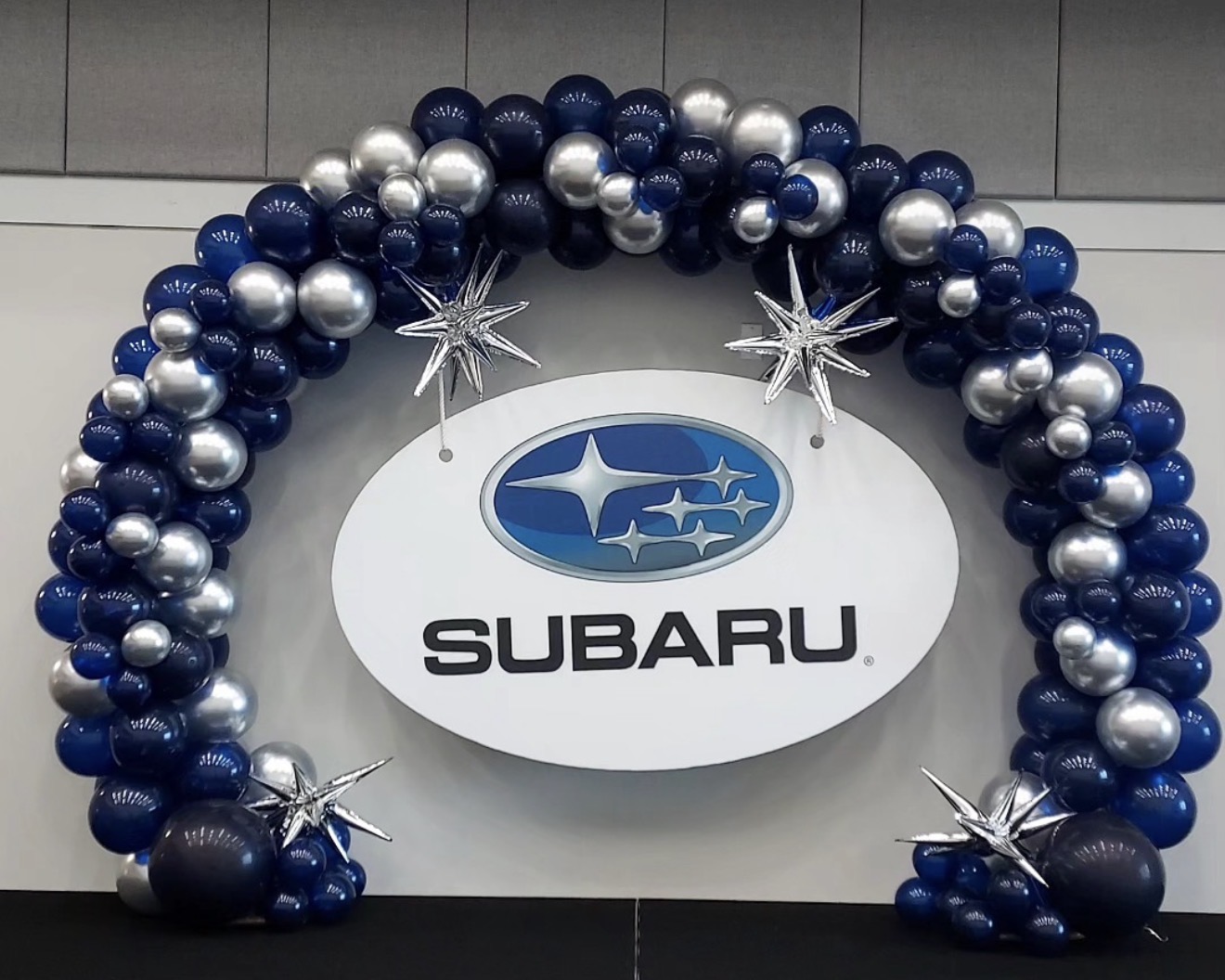 Decorative arch of blue, silver, and white balloons surrounds Subaru logo sign with stars and snowflake decorations.