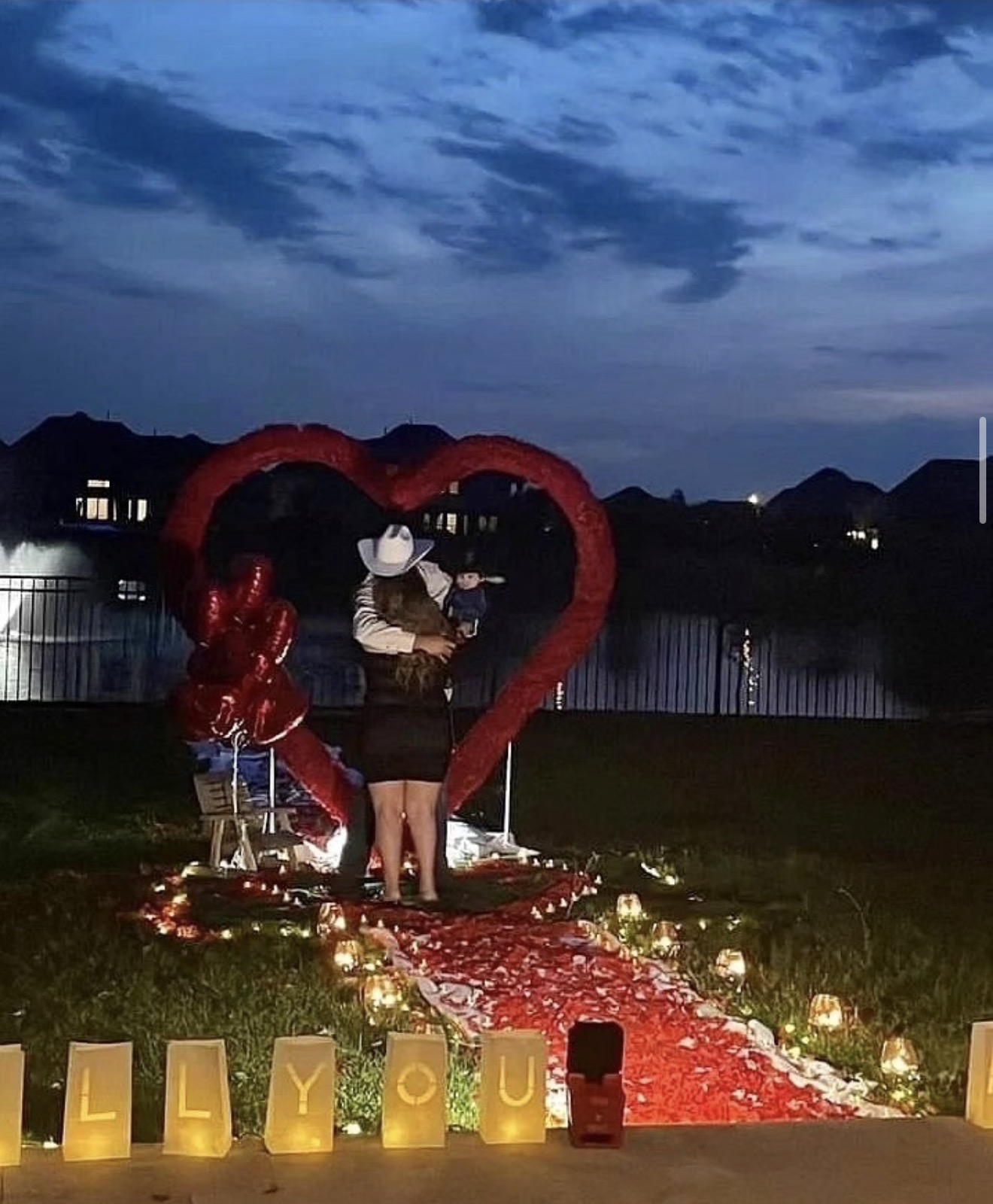 Two people stand under a heart-shaped balloon arch on a rose petal path at dusk, with a lake and mountains in background.