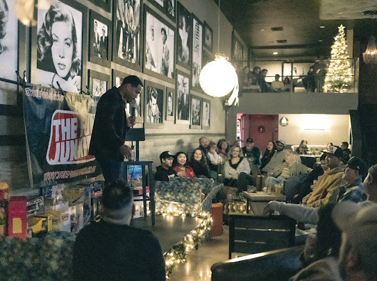 Man speaking to seated audience in a decorated room with framed photos, Christmas tree, and festive lights.