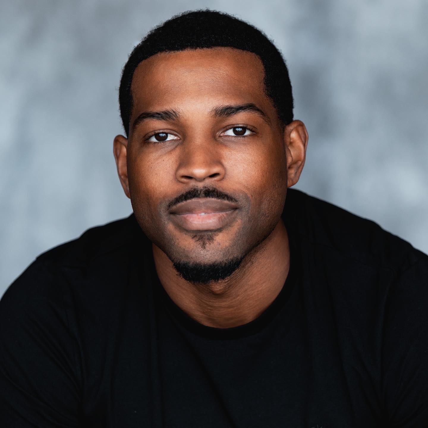 Portrait of a man with short hair and a slight smile, wearing a black shirt, against a gray background.