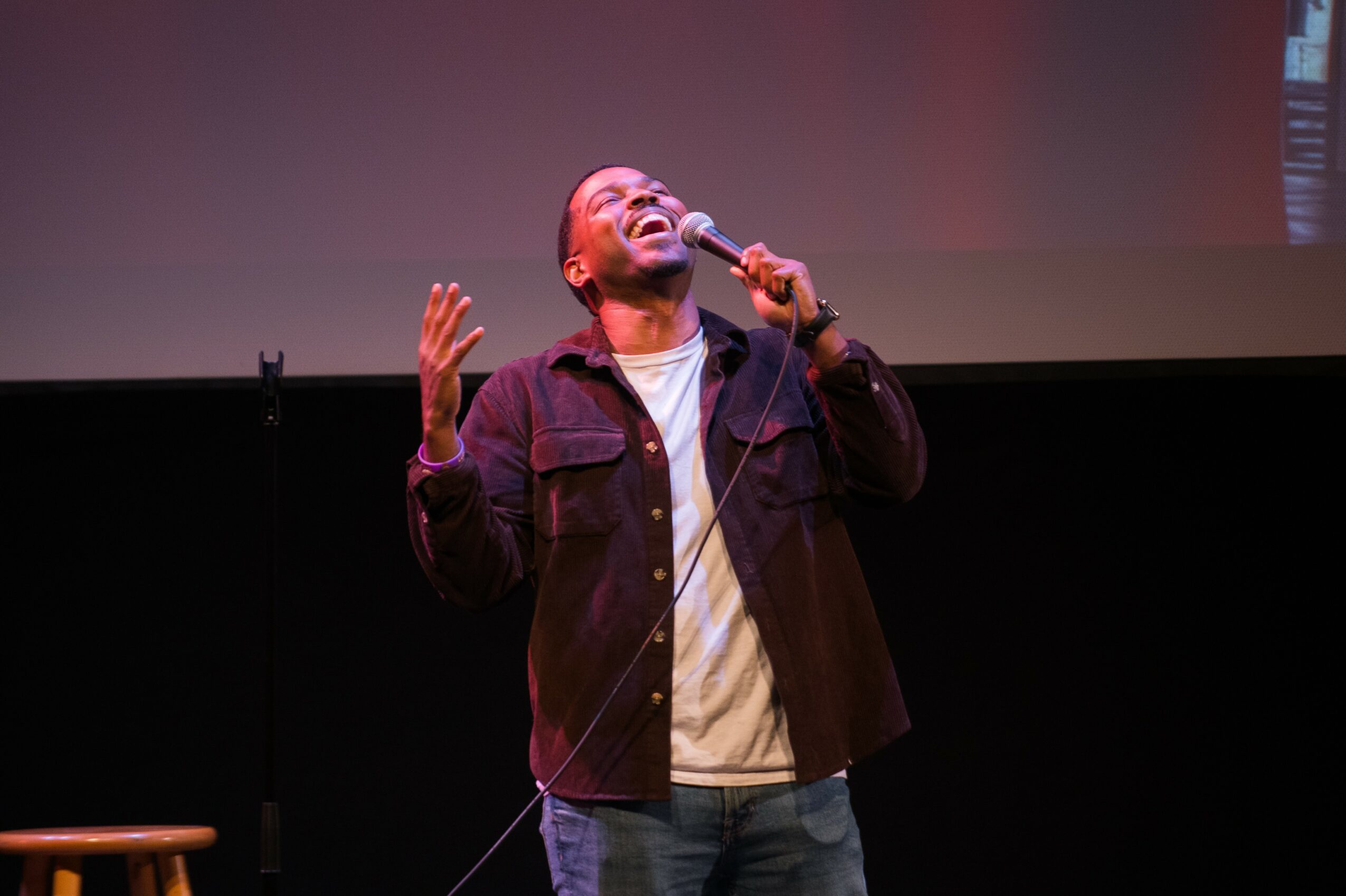 Person performing stand-up comedy on stage, holding microphone, with eyes closed and hands raised, in front of a large screen.