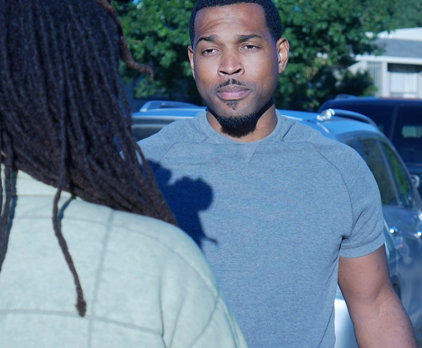 Two people talking outdoors, one facing the camera with a serious expression, the other with back to the camera, trees in background.