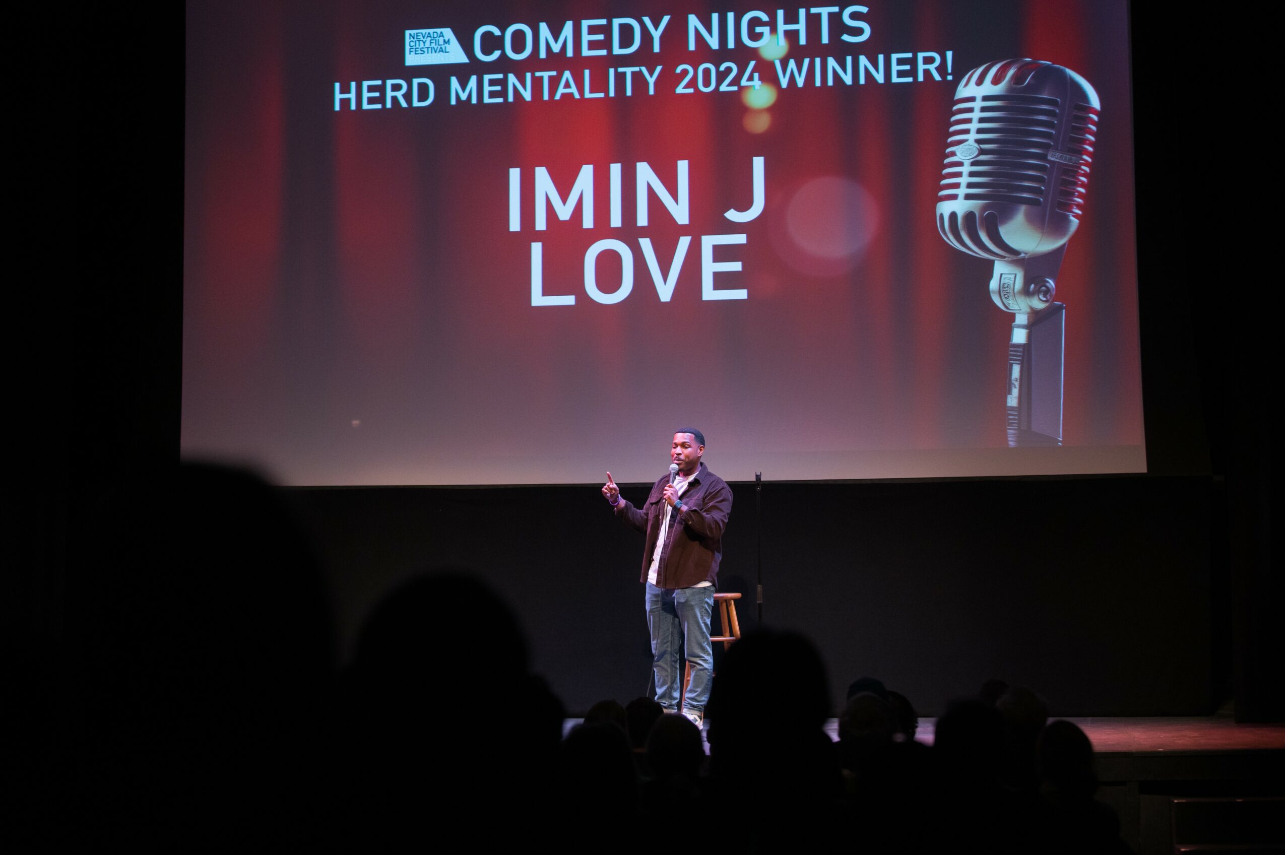 Person on stage speaking into a microphone at a comedy event, with a large screen behind displaying text and a microphone image.