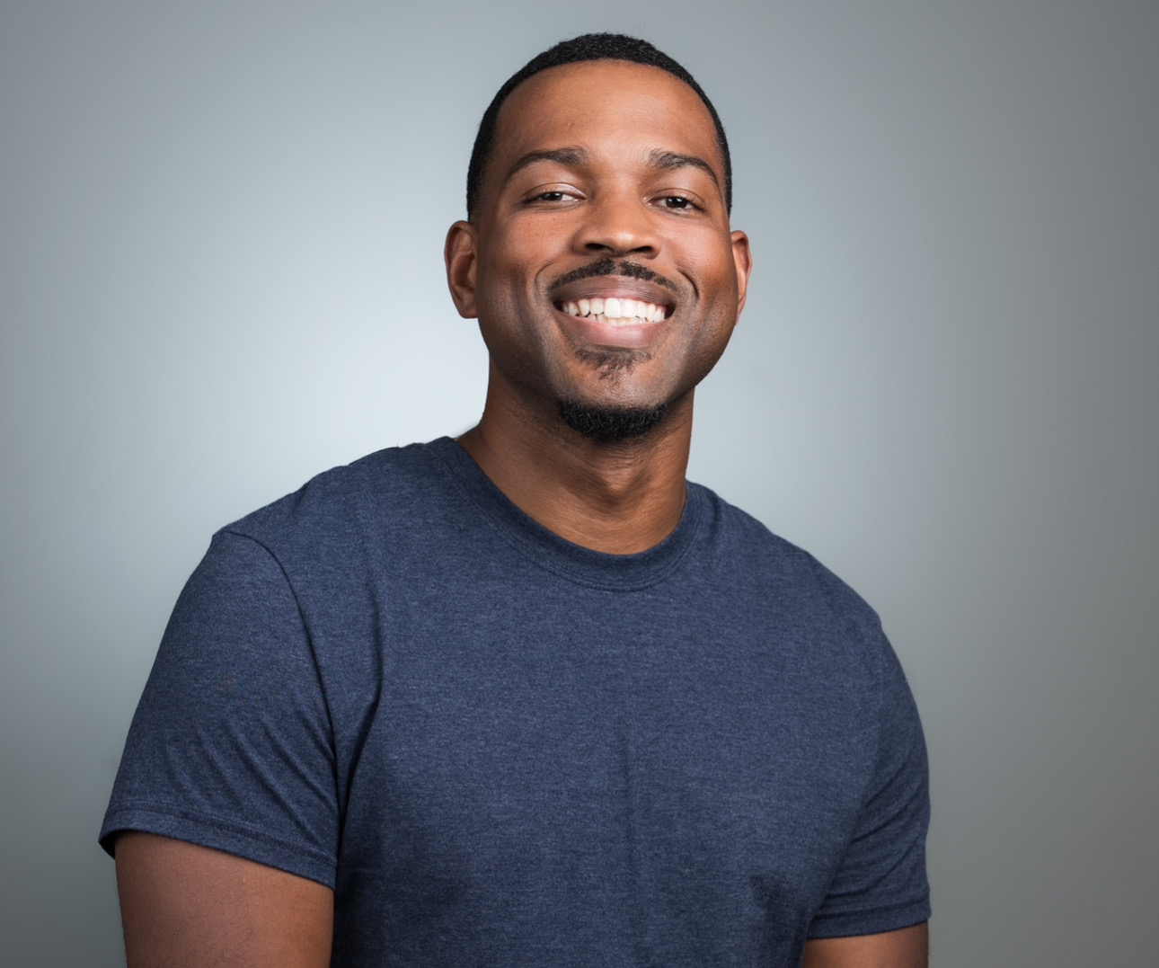 Smiling man with short hair and a beard wearing a dark blue t-shirt, facing forward, neutral background.