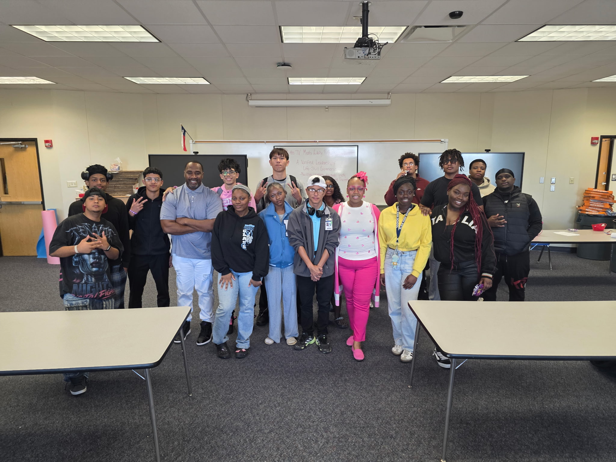 Group of students standing in a classroom, some smiling and waving, with desks and a whiteboard behind them.