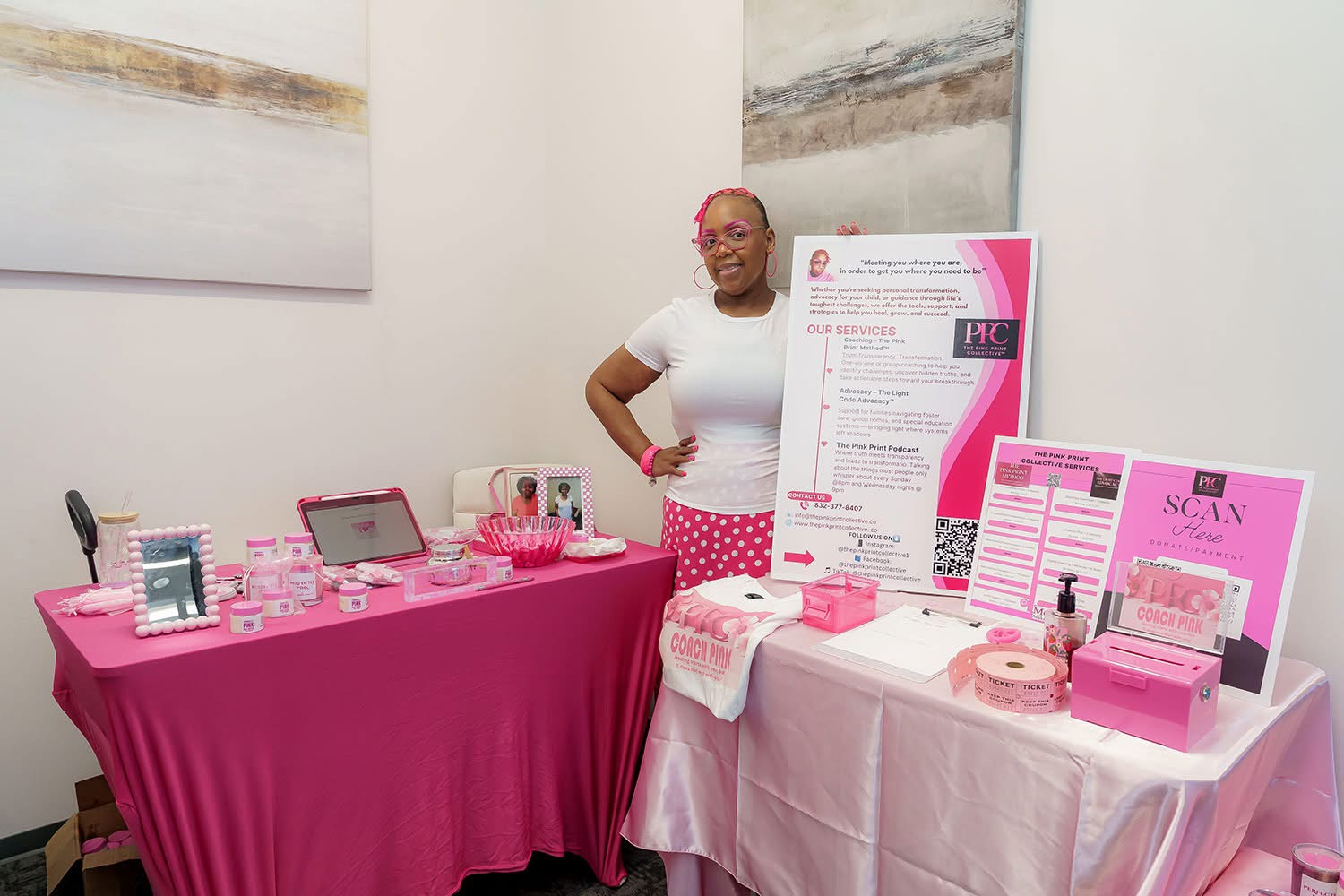 Woman standing next to a pink-themed display table with skincare products and informational posters, in a white room.