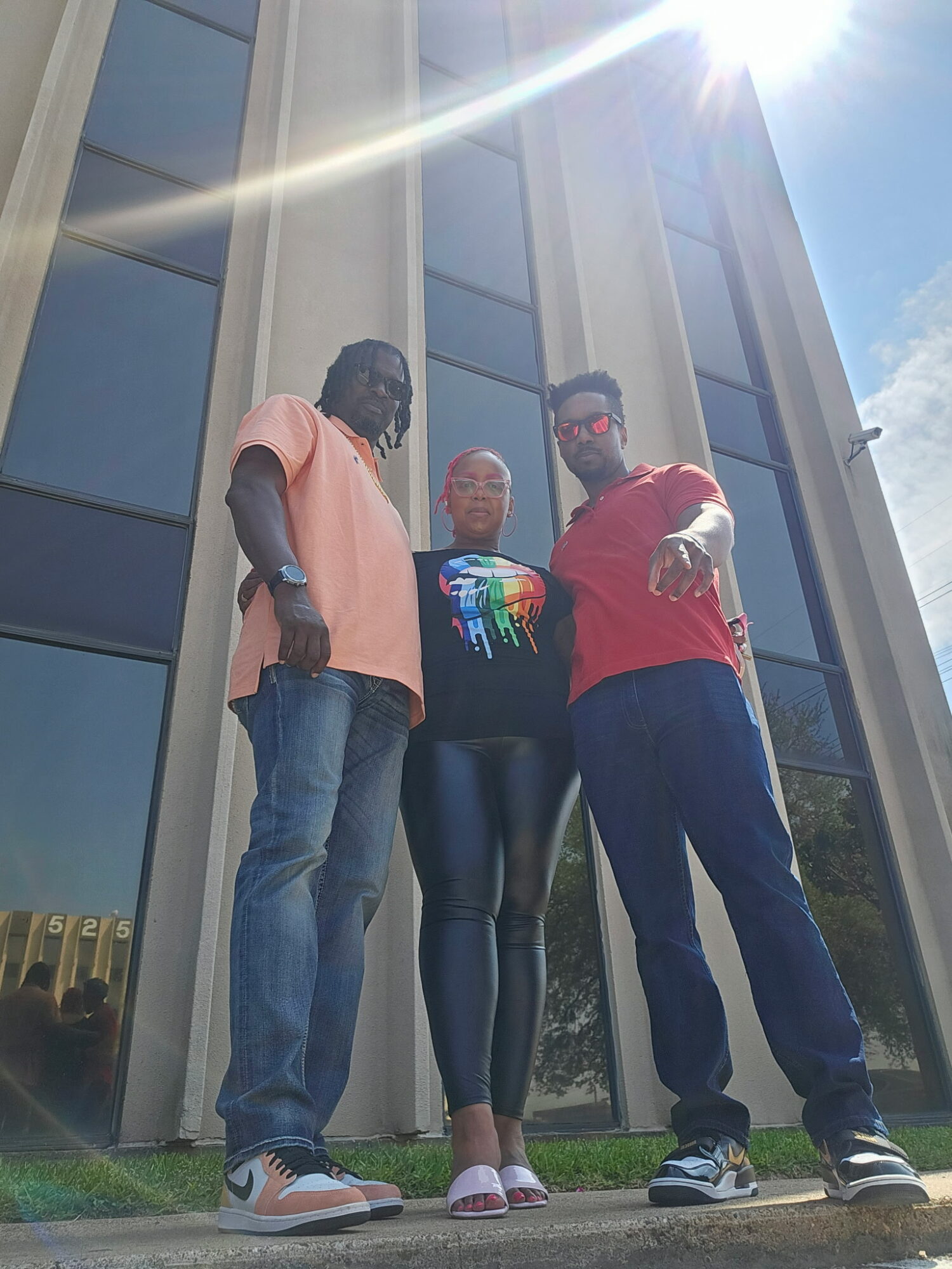 Three people standing outside a modern building with large glass windows, sunlight shining overhead.