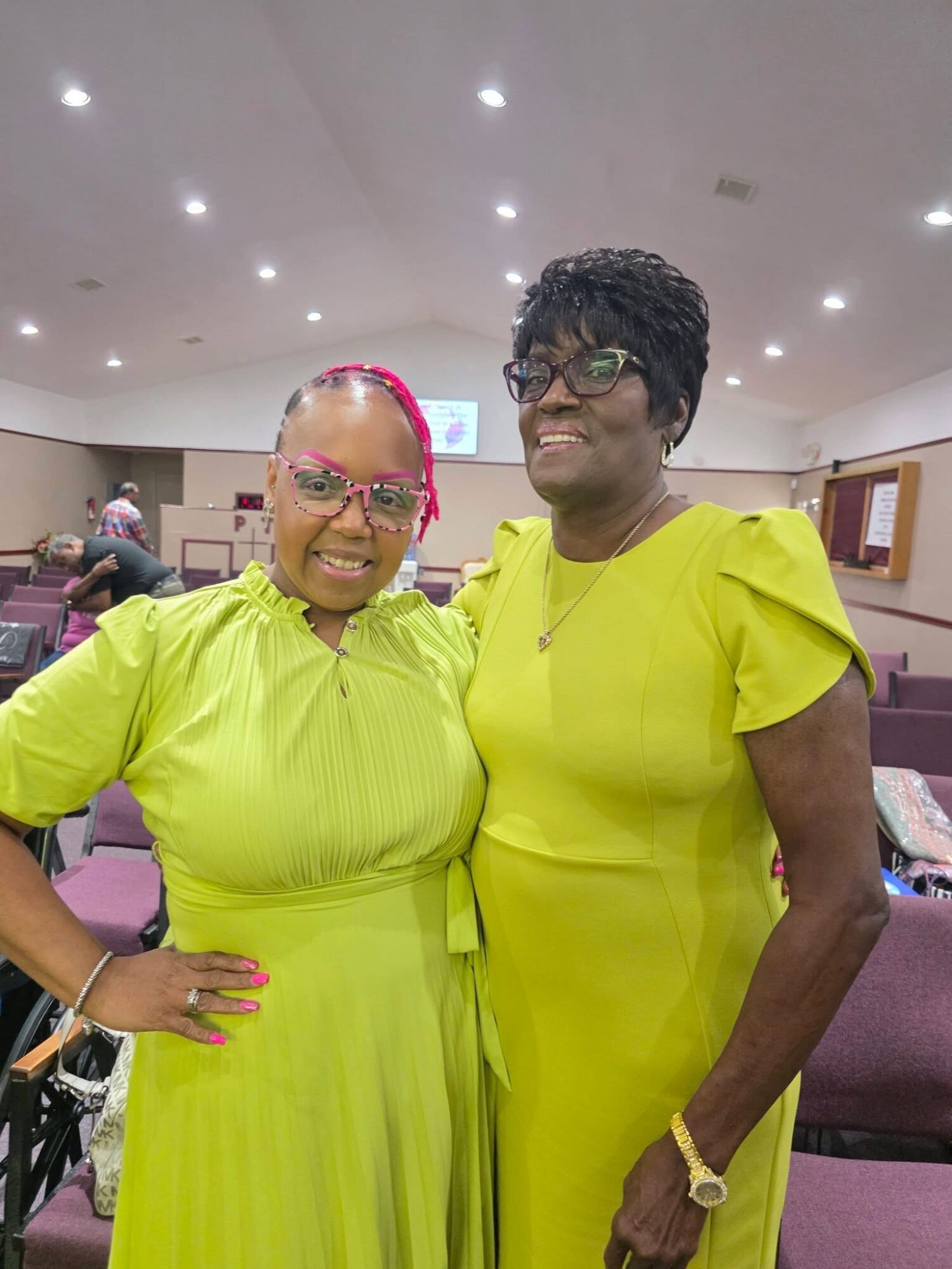 Two women stand together smiling in a room with purple chairs, wearing bright yellow dresses and glasses.