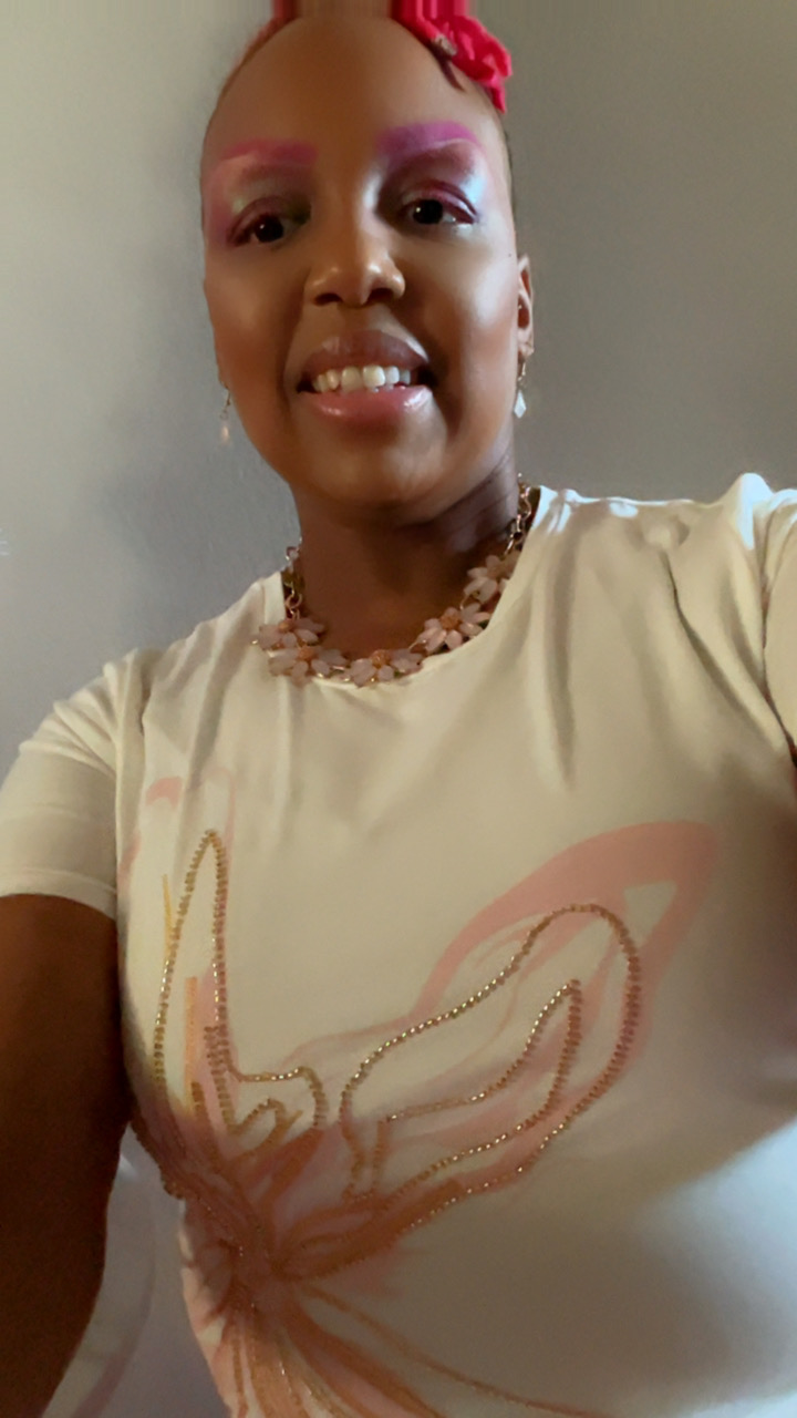 Woman smiling with pink makeup, red hair accessory, wearing a cream top and necklace, against a plain background.