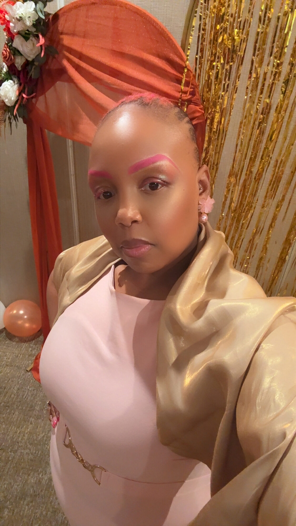 Person with makeup and a beige jacket taking a selfie at an event with gold and orange decorations.