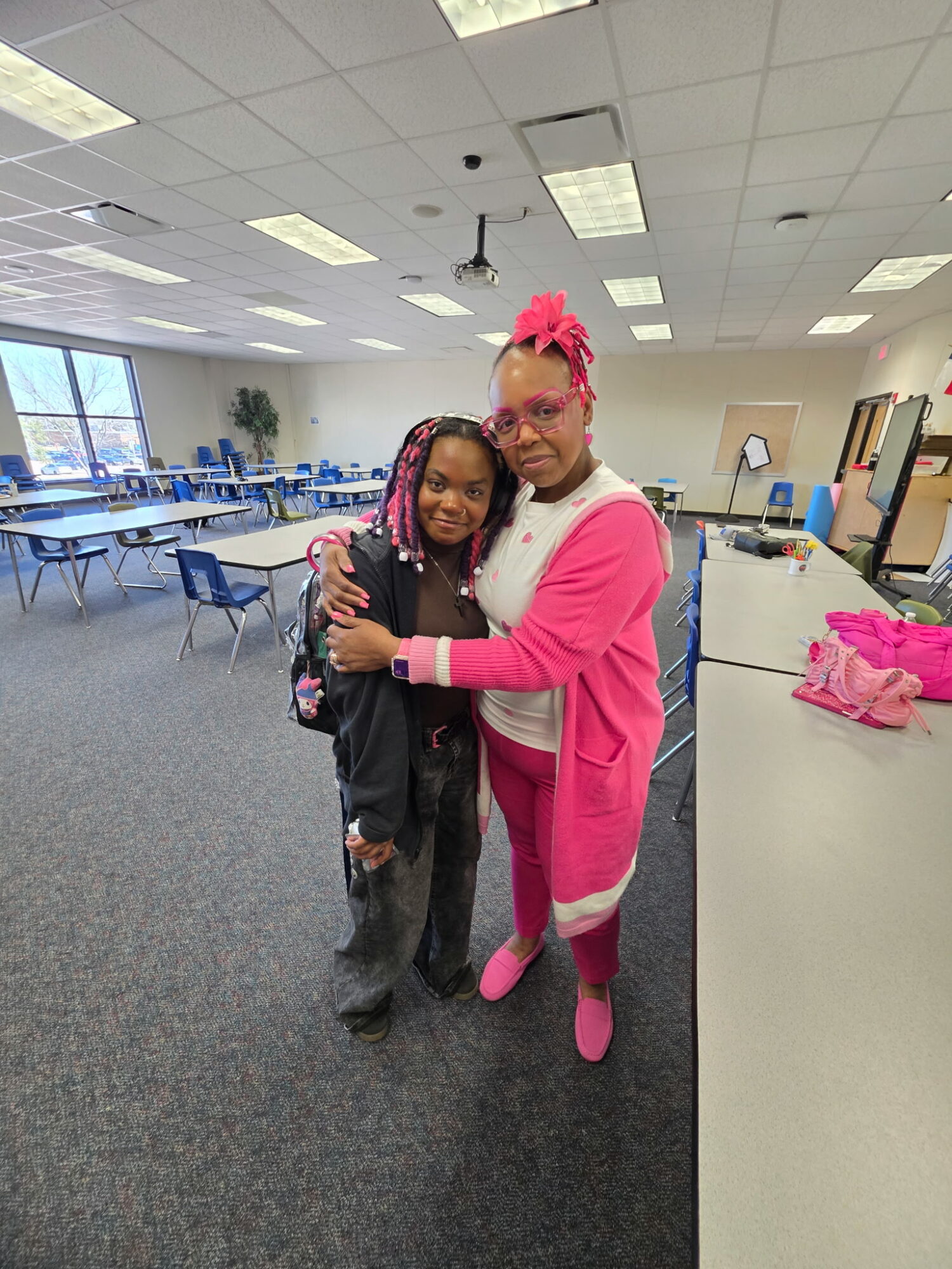 Two people hugging in a classroom, one with pink hair and glasses, the other with long hair, smiling.