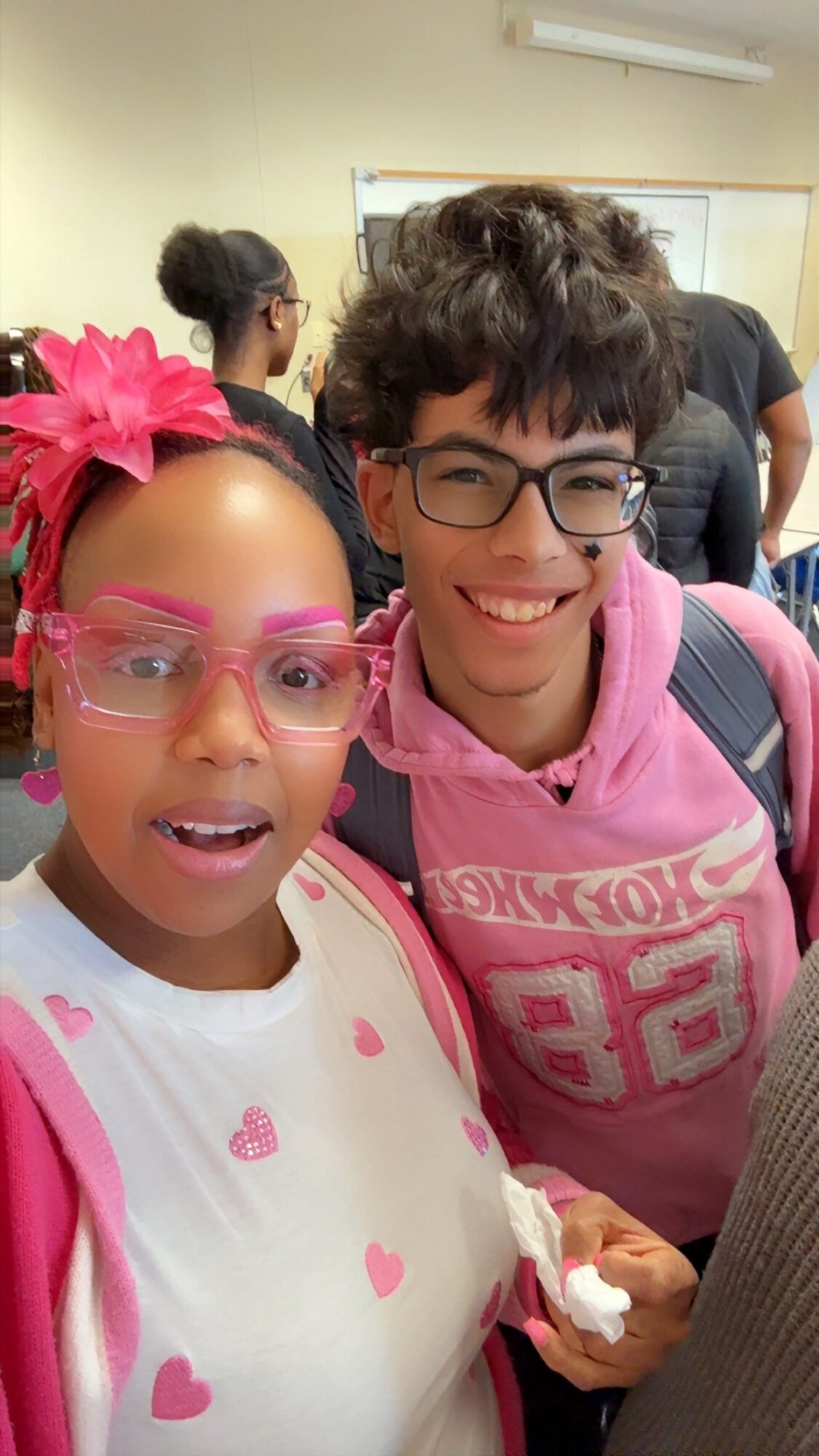 Two students smiling in a classroom, one wearing pink glasses and a pink bow, the other in a pink hoodie.