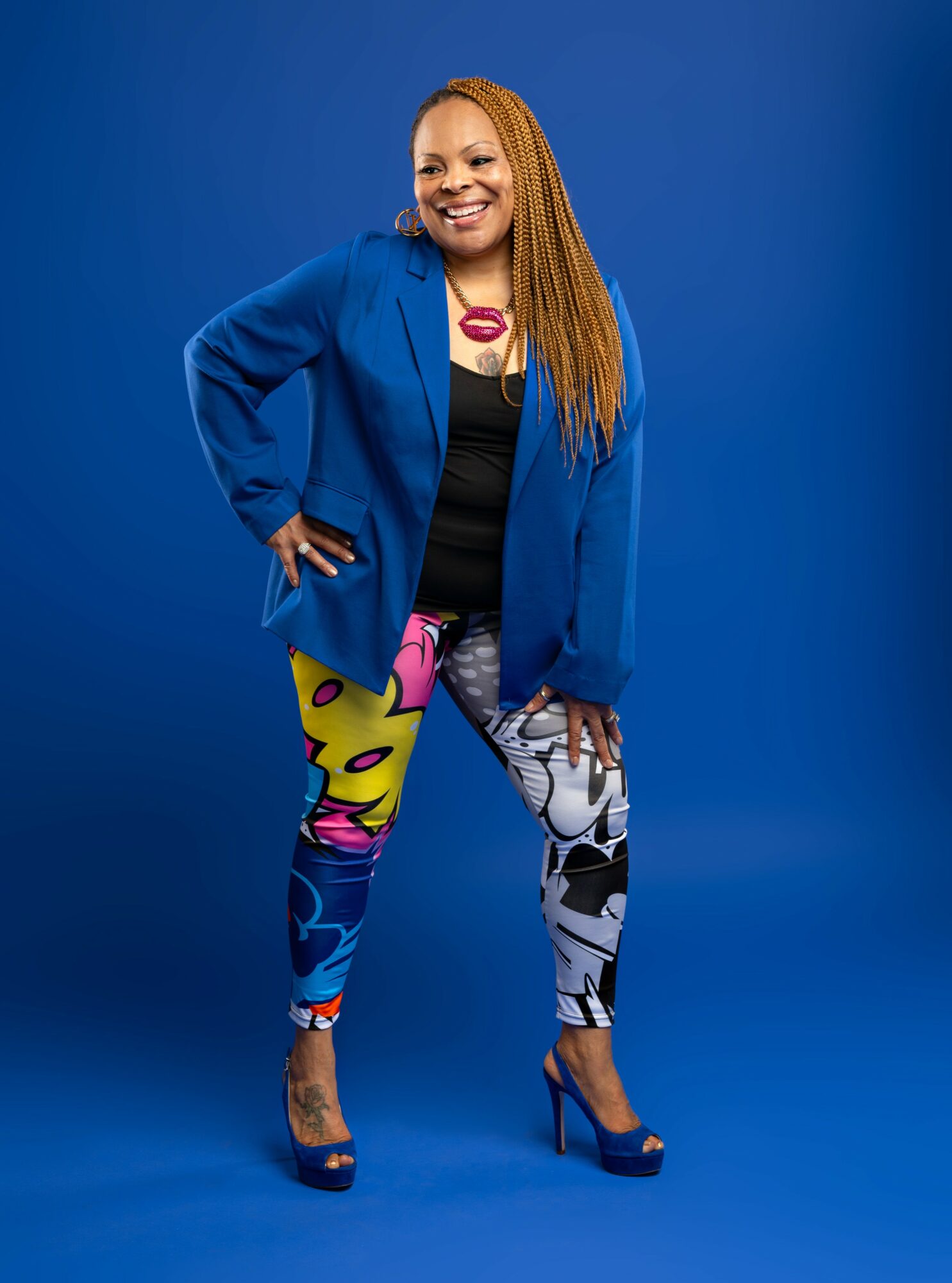 Woman with long hair in a blue blazer, colorful leggings, and high heels, smiling against a blue background.