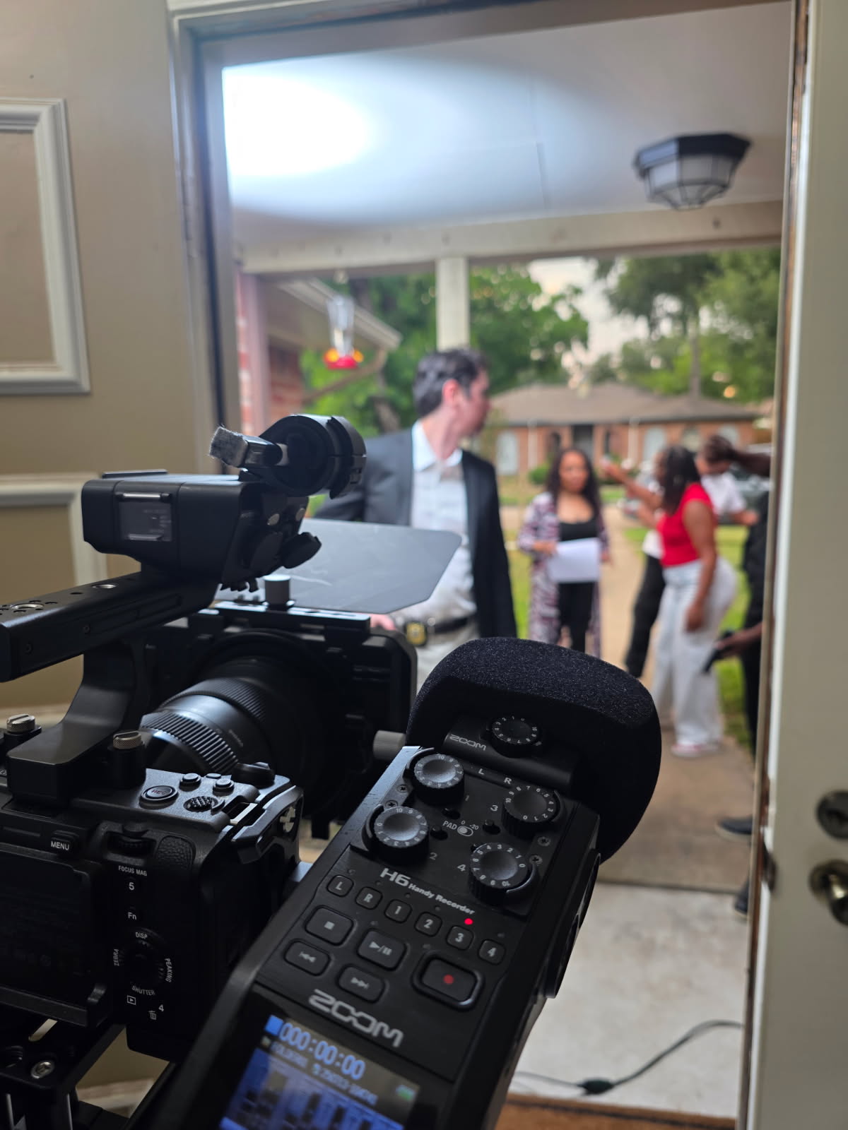 Camera inside a house capturing people outside through a window, with a person in a suit and others in casual clothing.